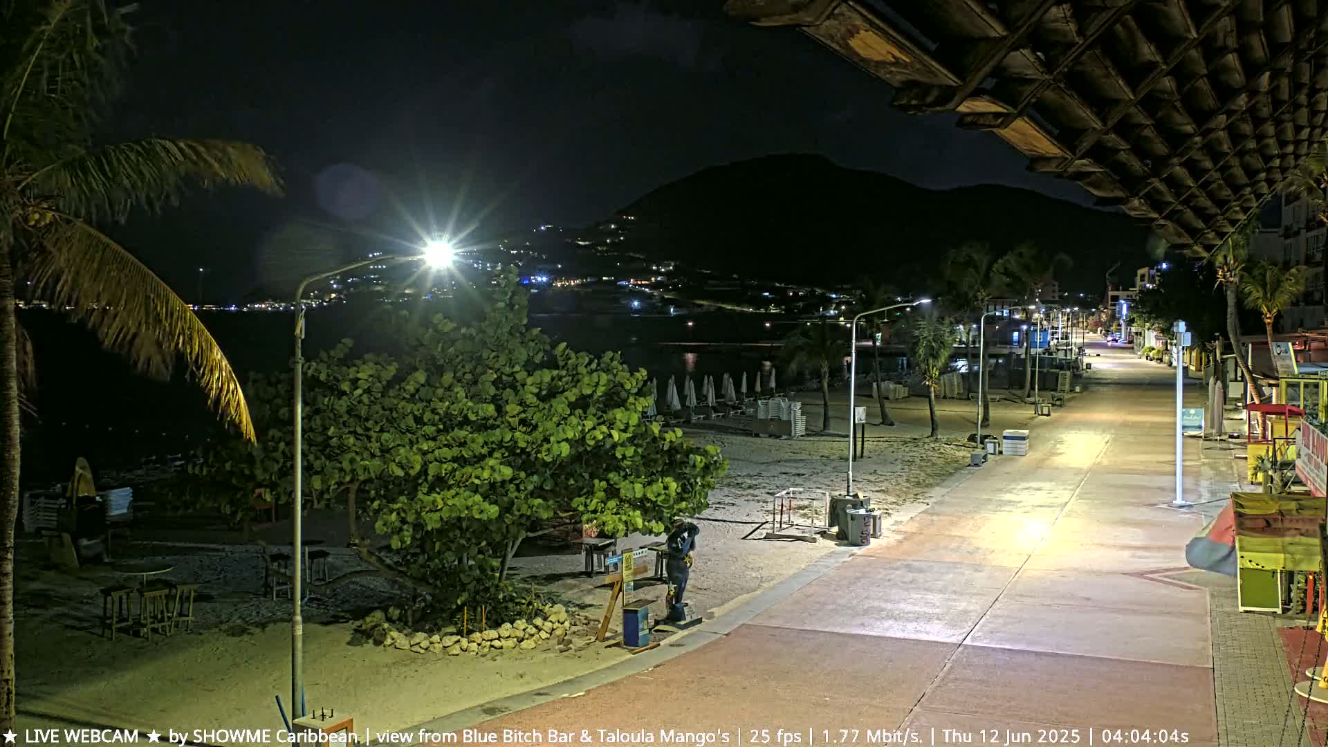 A nighttime beach scene shows a paved walkway leading to a dark ocean under a partly cloudy sky, with lights illuminating the path and a few buildings in the background.