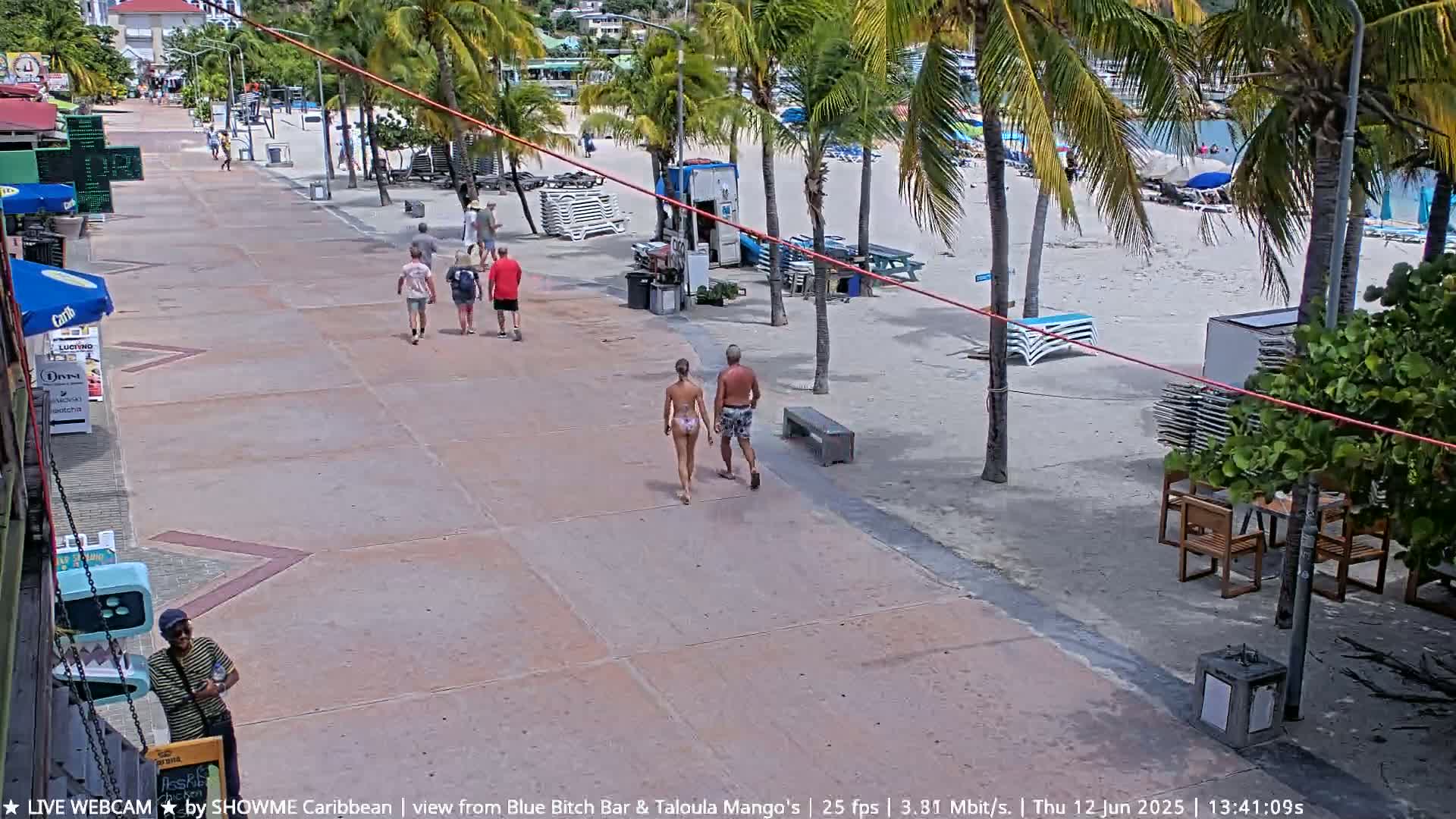 A paved walkway alongside a sandy beach lined with palm trees and beach chairs, with several people walking in different directions under a sunny sky.