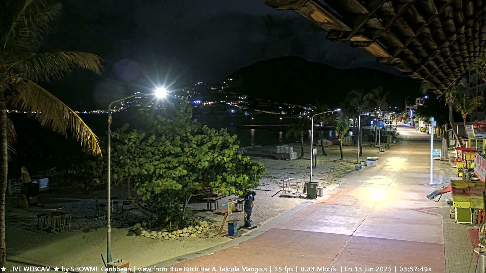 A nighttime beach boardwalk scene shows a mostly empty beach with a few lights illuminating the path and nearby buildings; a dark mountain is in the background.