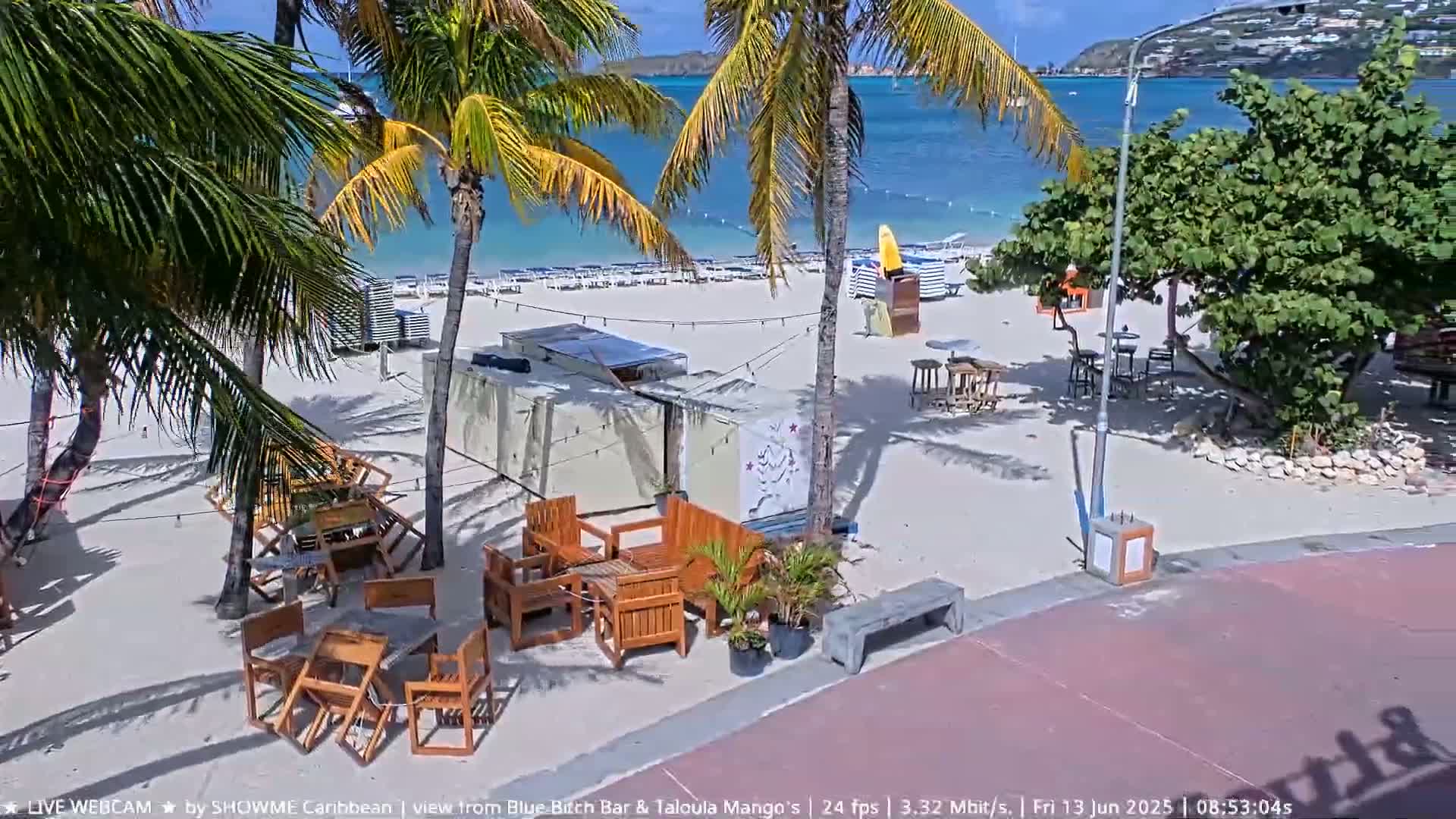 A sandy beach with palm trees, outdoor seating areas, and calm blue water under a sunny sky.
