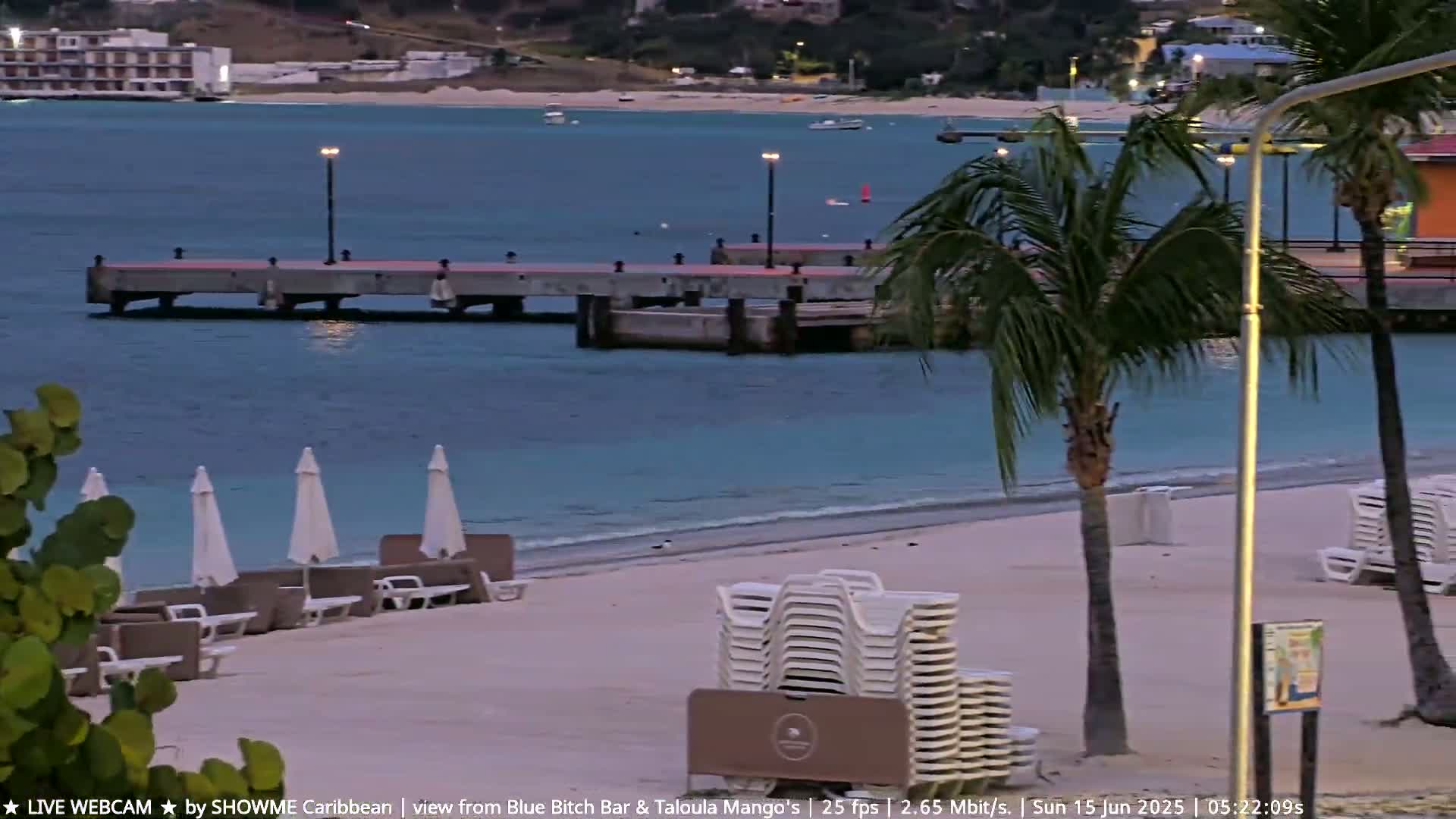A tranquil beach scene at dusk shows a pier extending into calm, turquoise water, with palm trees, beach chairs, and umbrellas on the sandy shore.