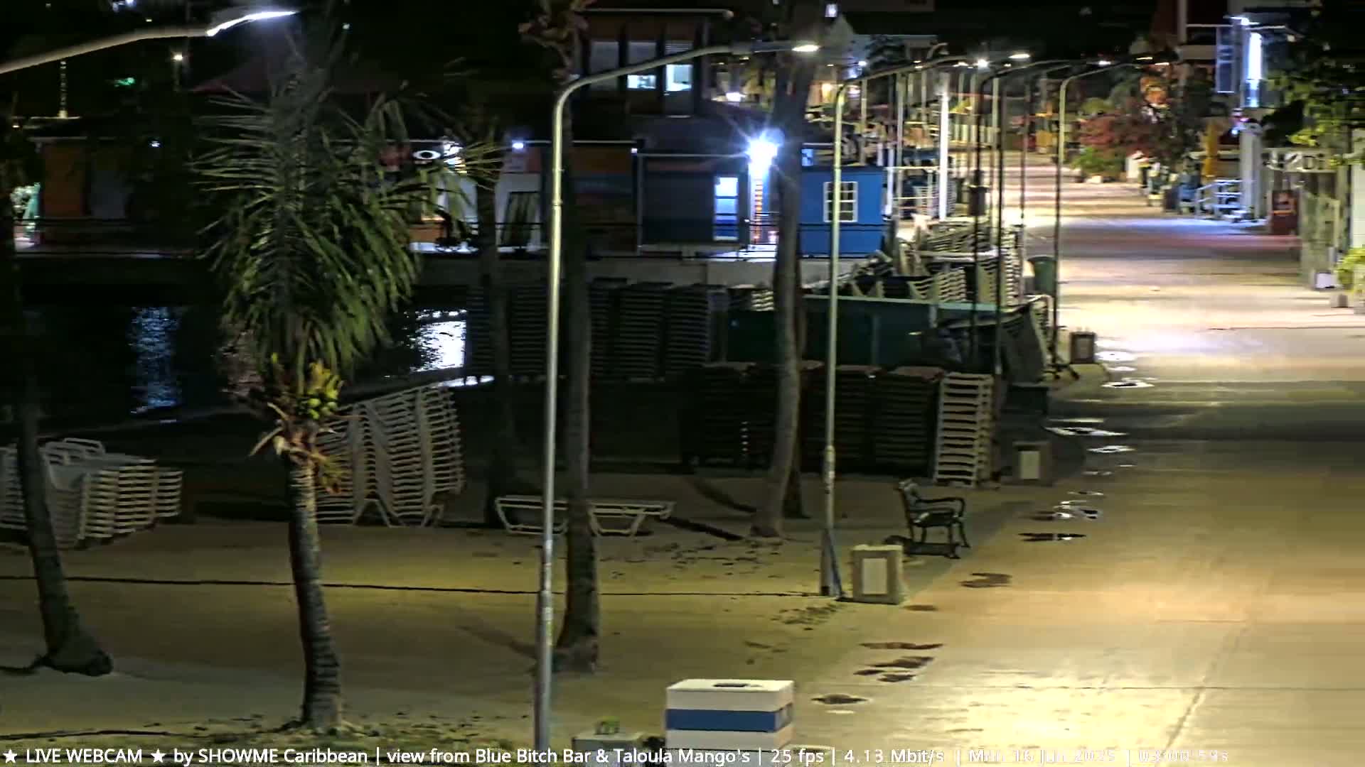 A nighttime view of a beachside walkway illuminated by streetlights, with stacked beach chairs and palm trees visible, and wet patches on the ground.