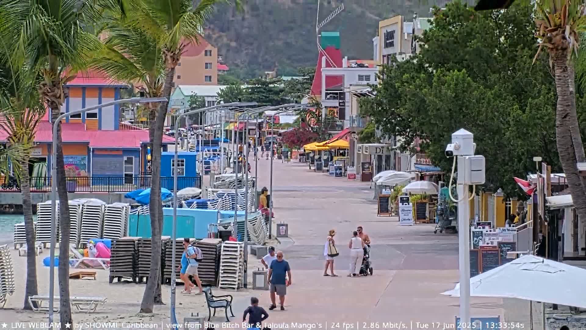 A mostly sunny waterfront boardwalk lined with colorful buildings and beach chairs, with a few people walking by.