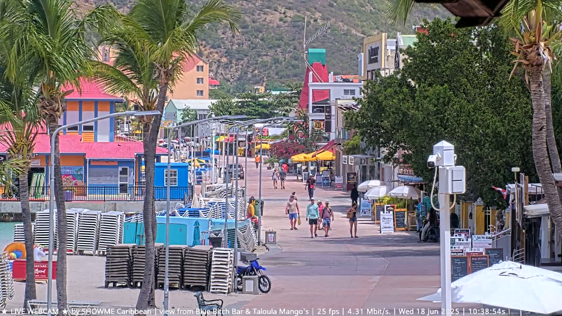 A sunny boardwalk scene shows pedestrians strolling past colorful buildings and beachside shops, with stacks of beach chairs lining the sand.