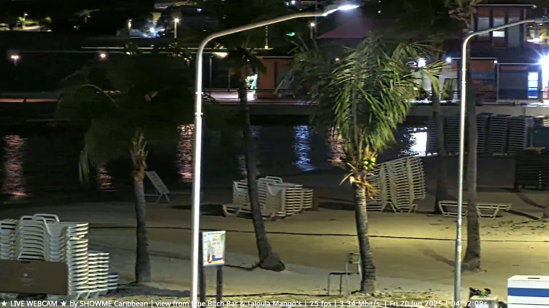 A nighttime beach scene shows calm water, stacked beach chairs, palm trees swaying gently in the breeze, and some buildings in the background.
