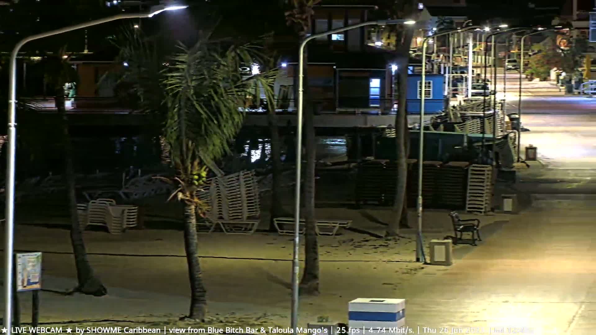 A nighttime beach scene shows stacks of beach chairs near palm trees and streetlights lining a paved walkway next to a building.