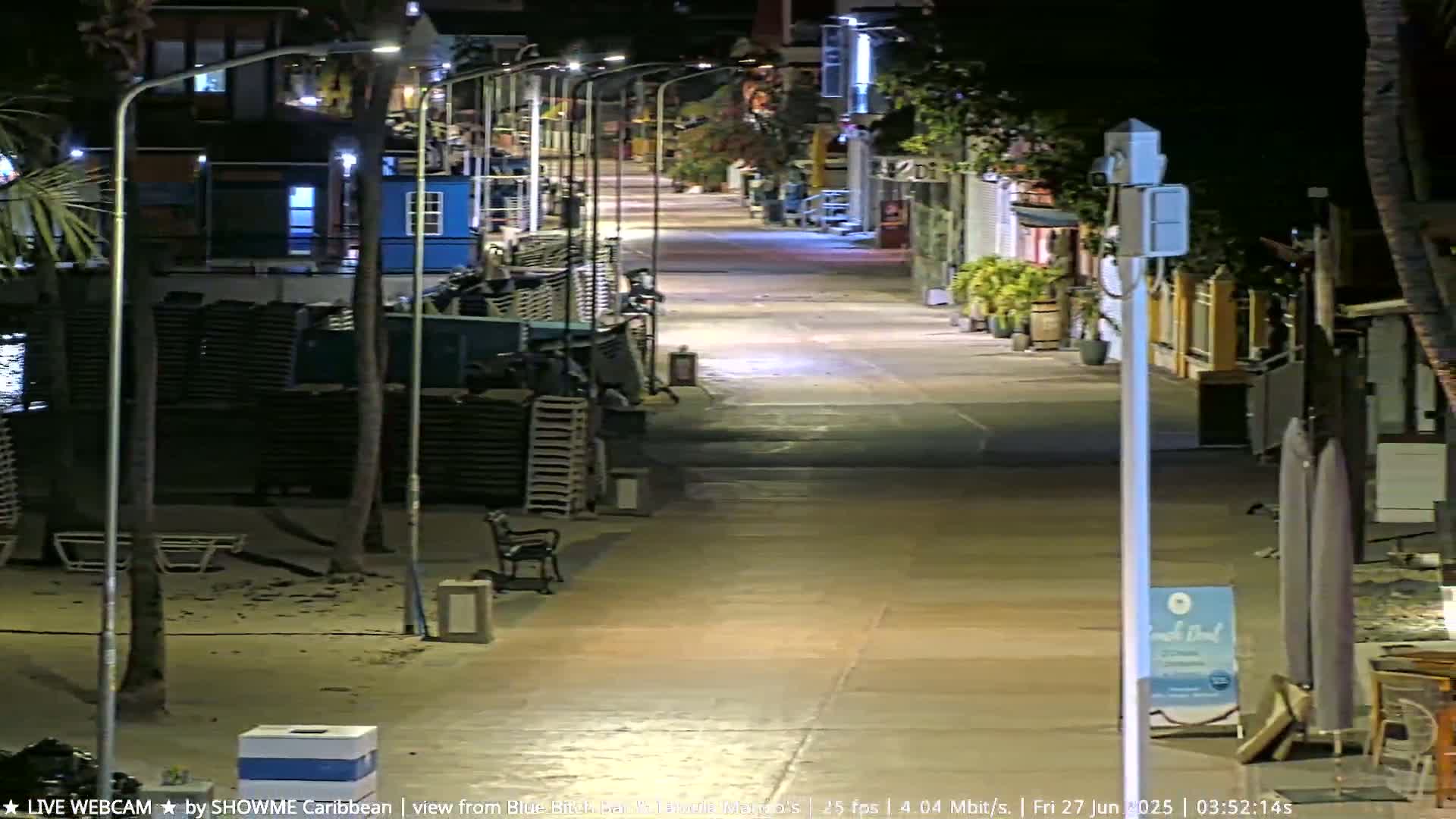 A deserted, brightly lit boardwalk at night, lined with closed shops and stacked beach chairs, under a clear sky.