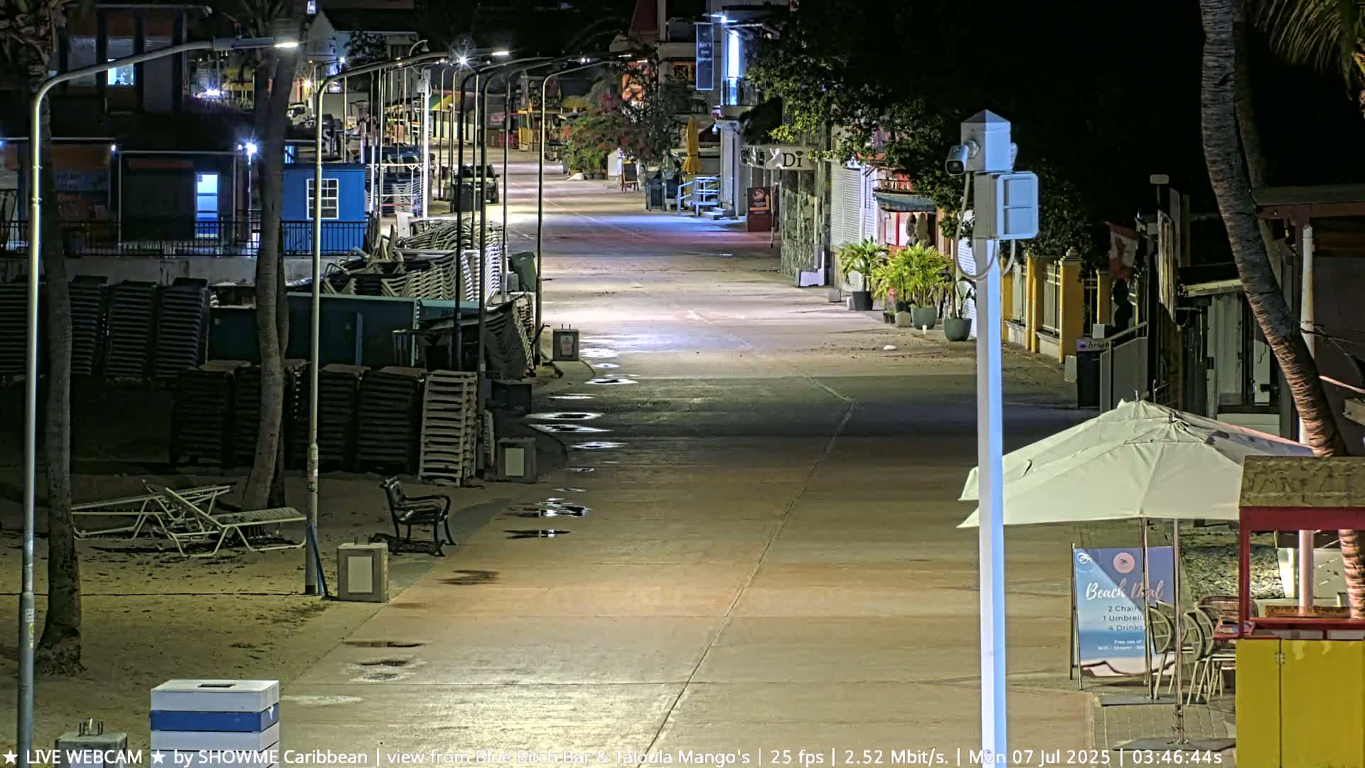 A nighttime, deserted boardwalk with stacked beach chairs and scattered puddles, lined with closed shops and palm trees under lamplight.