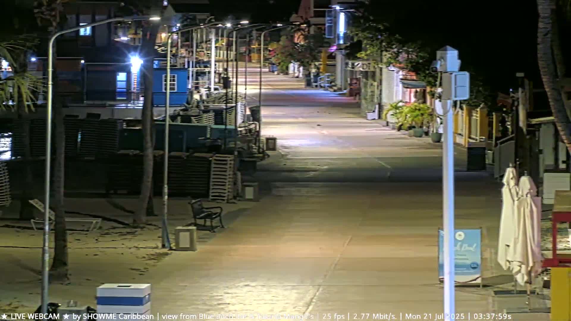 A nighttime, deserted beachfront promenade is illuminated by streetlights, with some beach chairs and closed beach umbrellas visible.