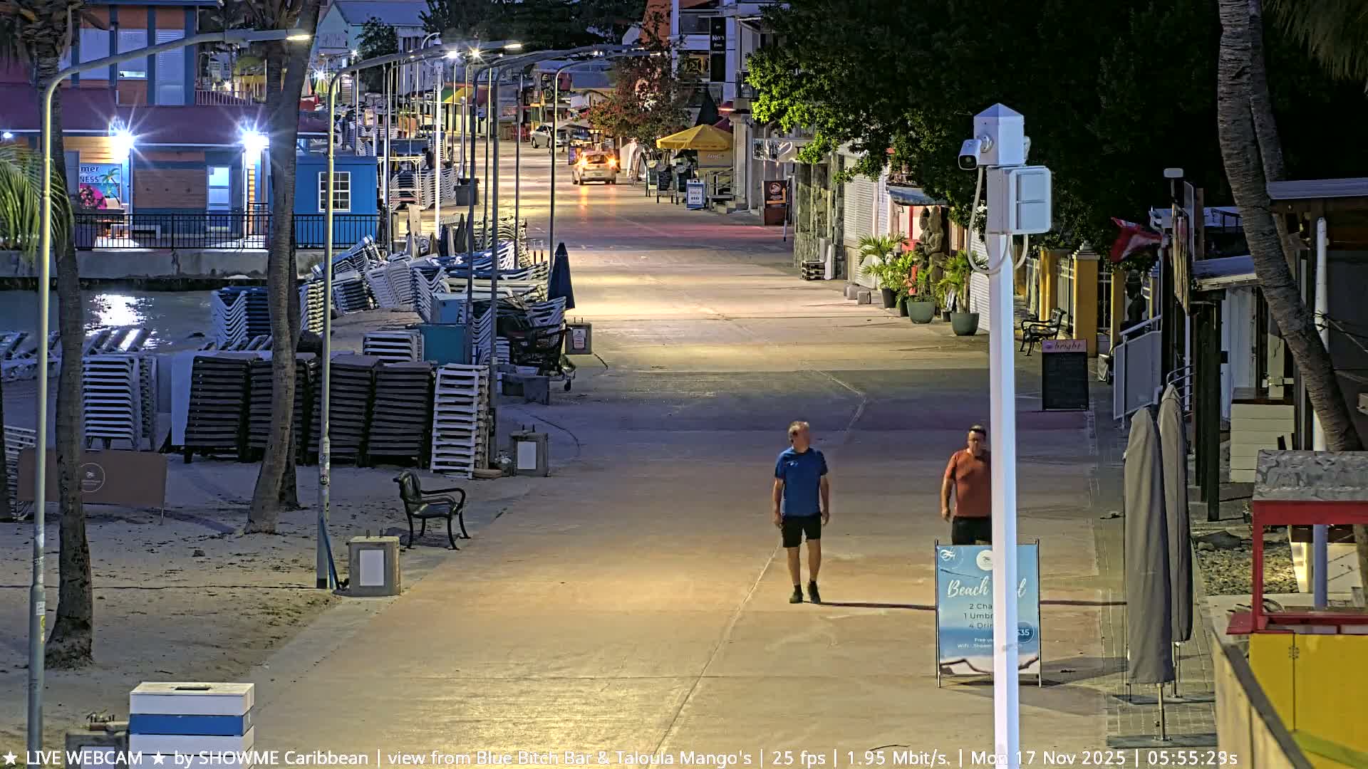 Two men walk along a brightly lit beach promenade lined with stacked chairs and illuminated buildings under a clear night sky.