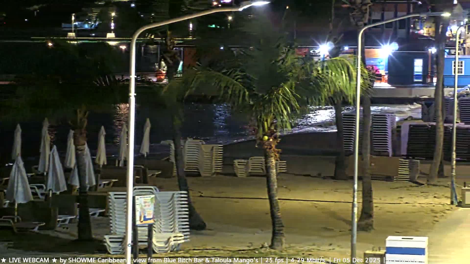 The image shows a tropical beach at night under clear skies, illuminated by artificial lights, with palm trees, stacked lounge chairs, and folded beach umbrellas along the water's edge.