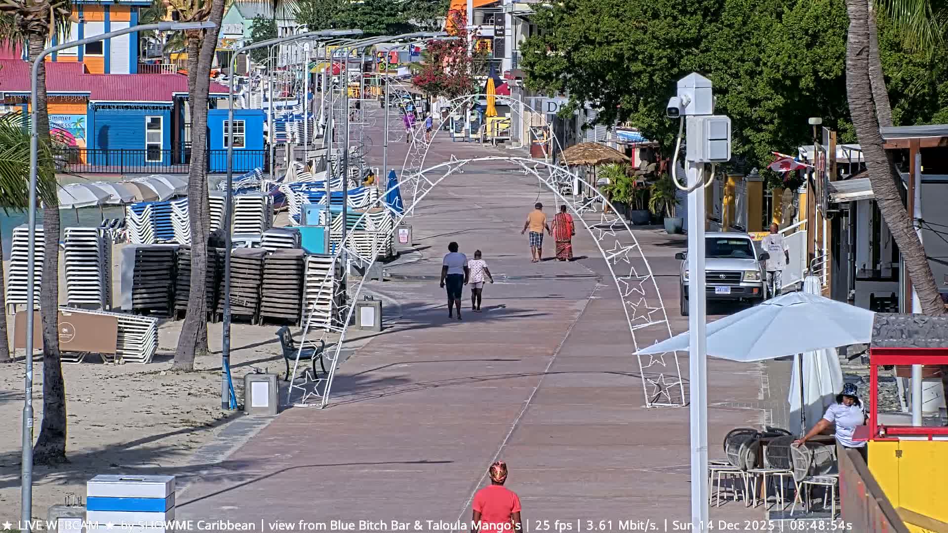 The image shows a tropical beach at night under clear skies, illuminated by artificial lights, with palm trees, stacked lounge chairs, and folded beach umbrellas along the water's edge.