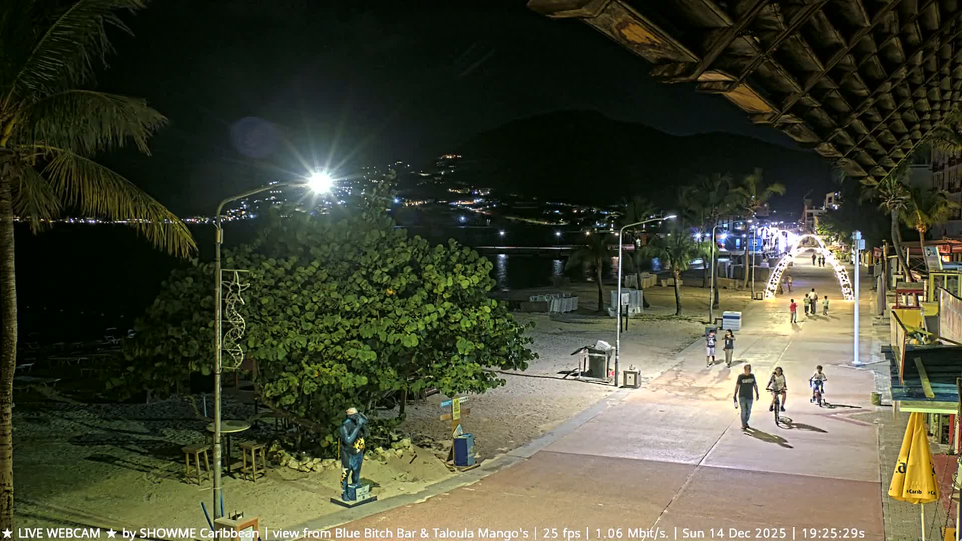 The image shows a tropical beach at night under clear skies, illuminated by artificial lights, with palm trees, stacked lounge chairs, and folded beach umbrellas along the water's edge.