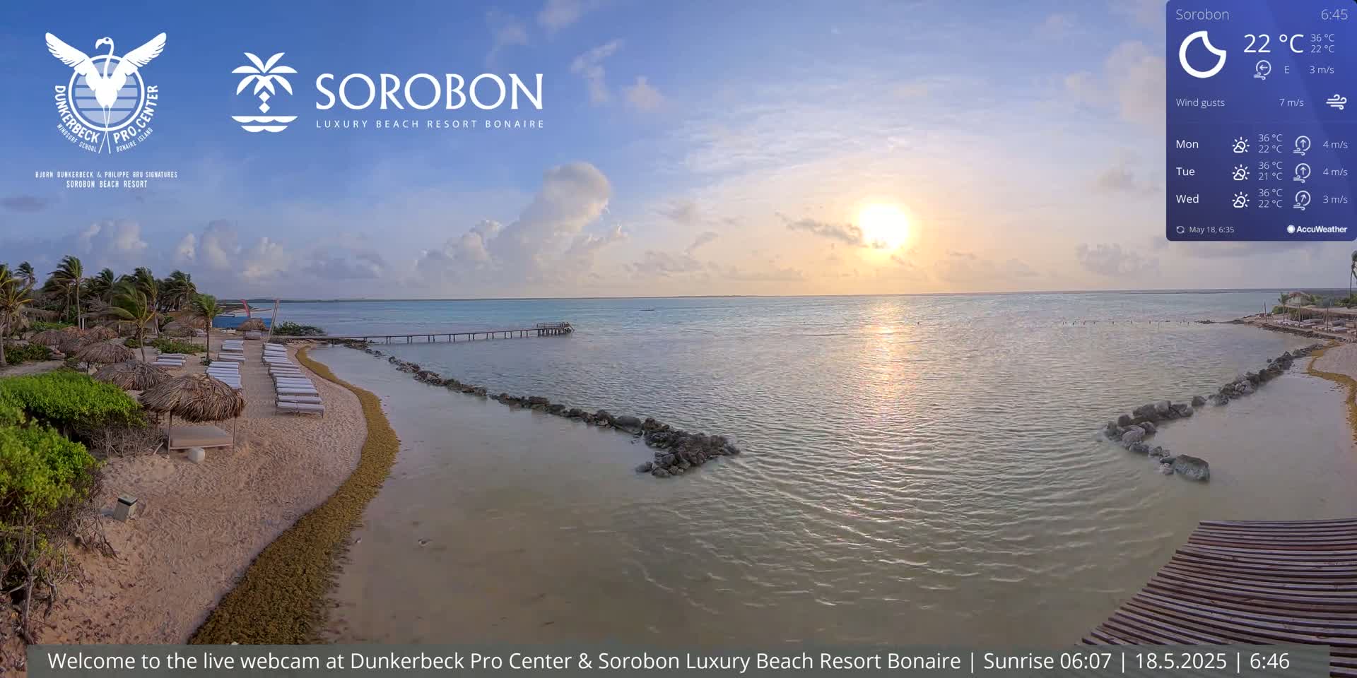 A beach with lounge chairs under thatched umbrellas at sunset, a calm ocean, and a pier extending into the water.