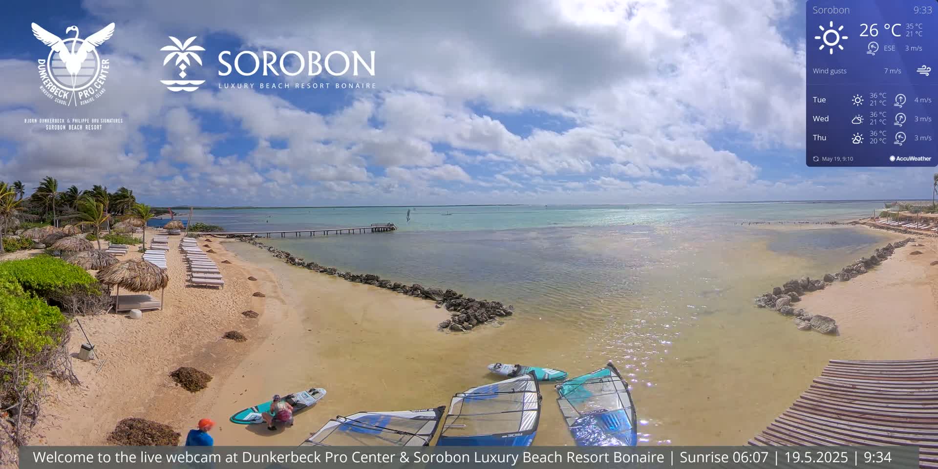 A sandy beach with lounge chairs and windsurfing equipment, under a partly cloudy sky, next to calm, shallow turquoise water.