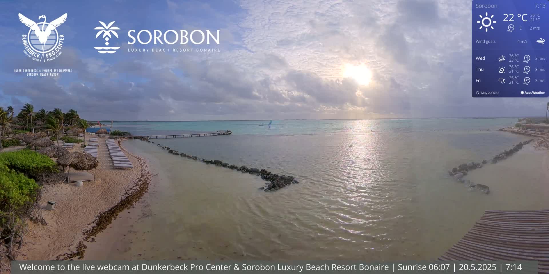 A beach with lounge chairs and thatch umbrellas sits on a calm, shallow body of water under a partly cloudy sky with the sun shining.