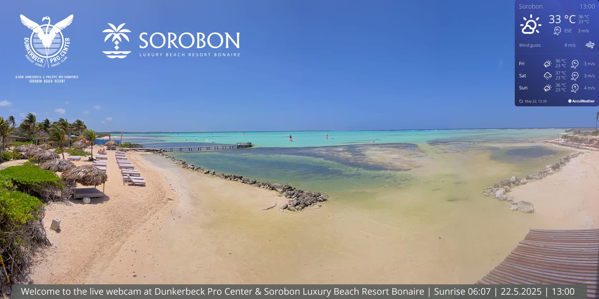 A sandy beach with lounge chairs and thatched umbrellas stretches toward a calm, shallow turquoise ocean under a bright, sunny sky.
