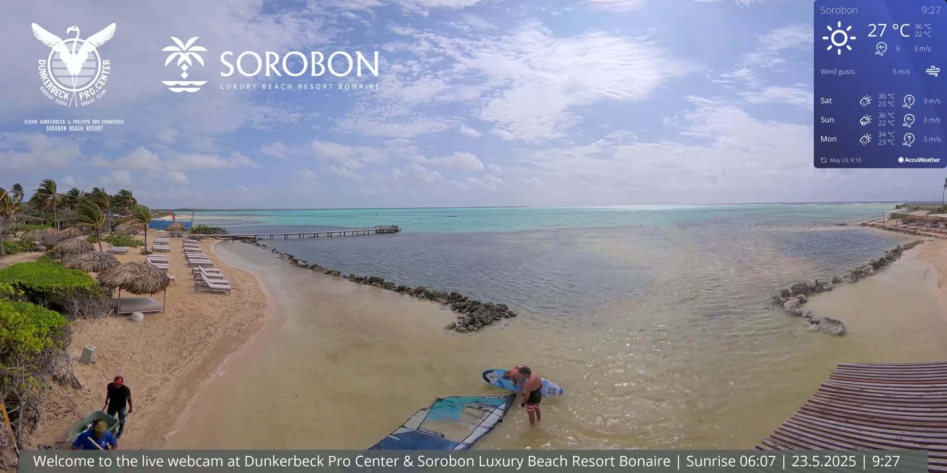 A sandy beach with lounge chairs and thatch umbrellas leads to calm, turquoise water under a mostly sunny sky.