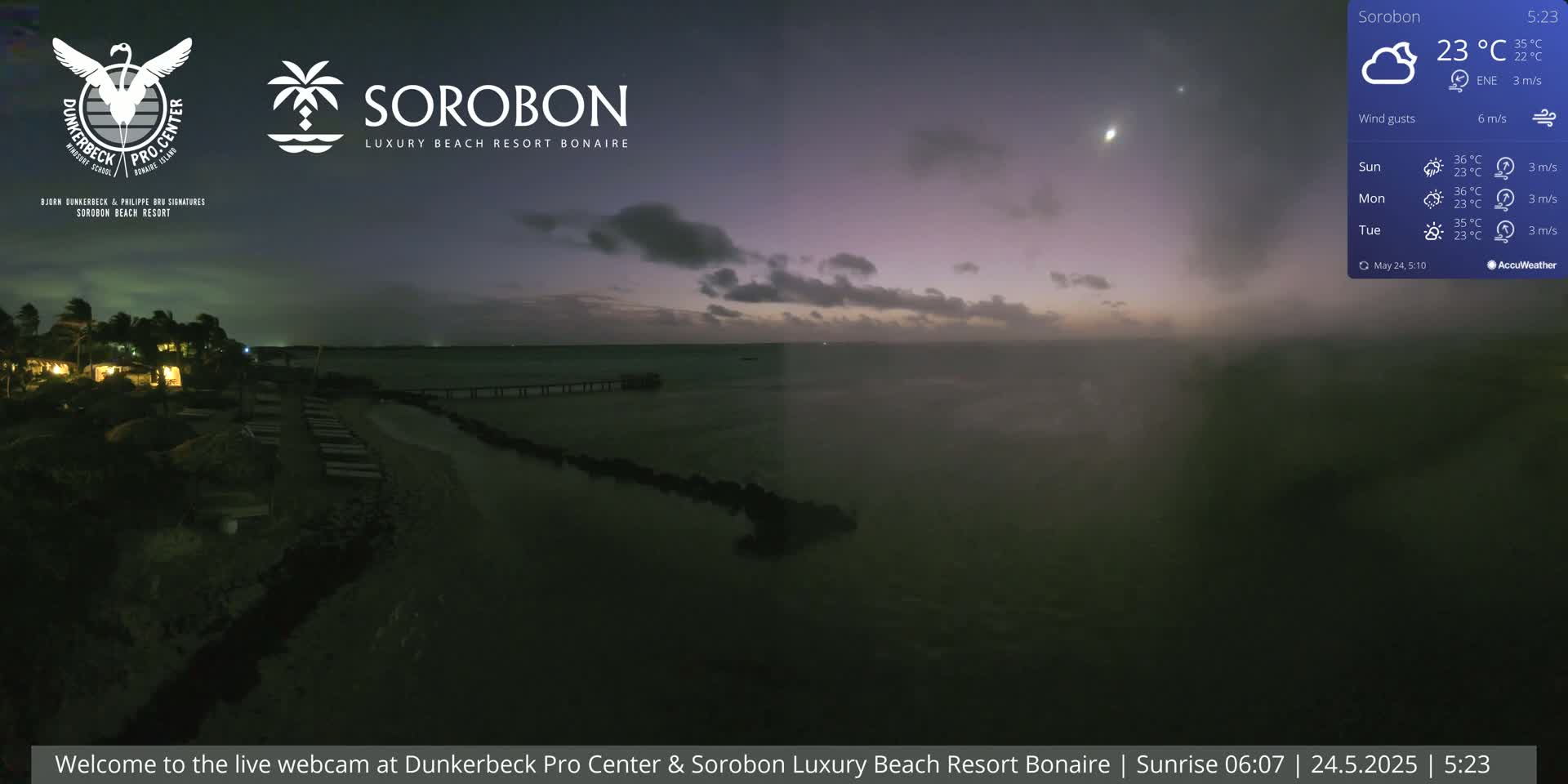 A nighttime beach scene shows a calm body of water under a mostly clear, dusky sky with a few clouds.