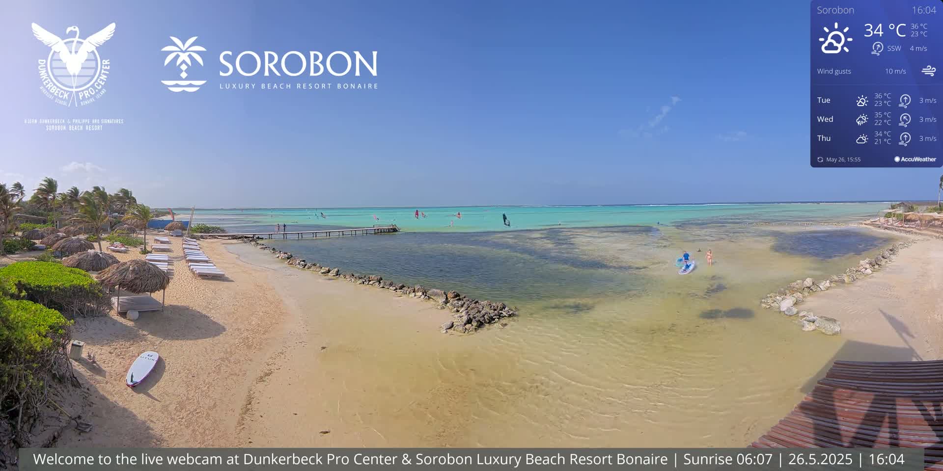 A sandy beach with lounge chairs and thatched umbrellas sits adjacent to calm, shallow turquoise water where windsurfers are visible on a sunny day.