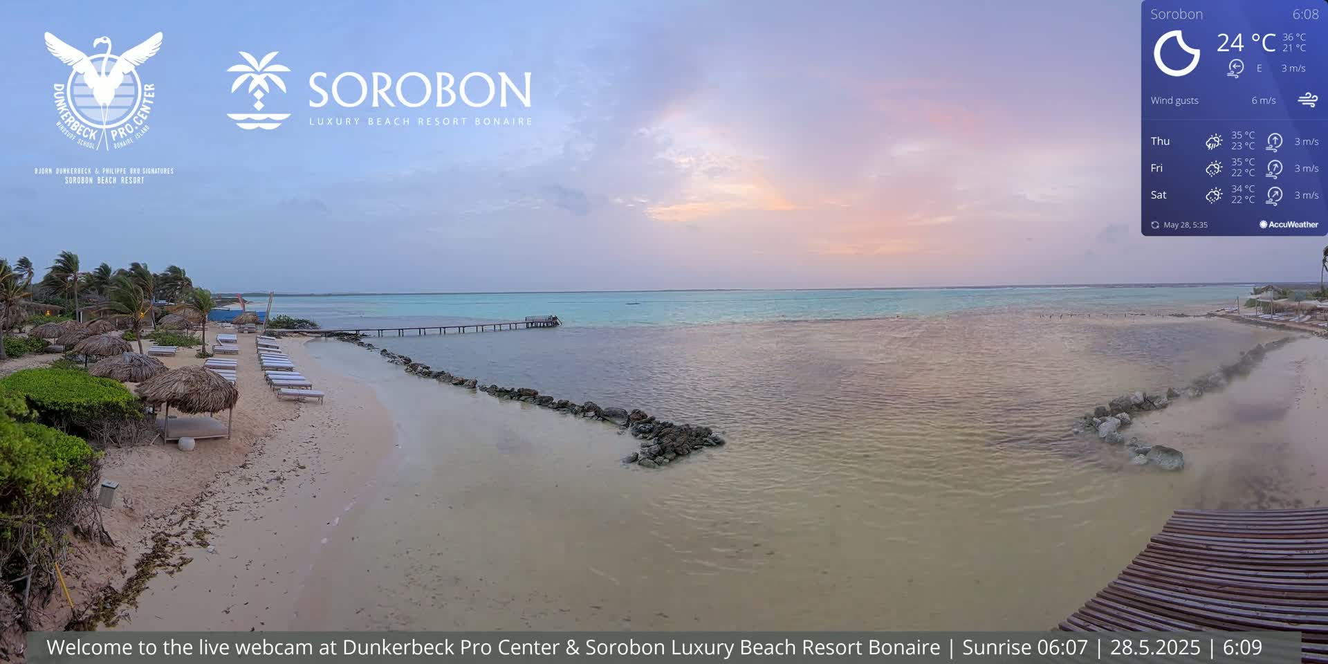 A tranquil beach scene at sunset shows calm, shallow water lapping a sandy shore lined with lounge chairs and thatch umbrellas under a pastel sky.