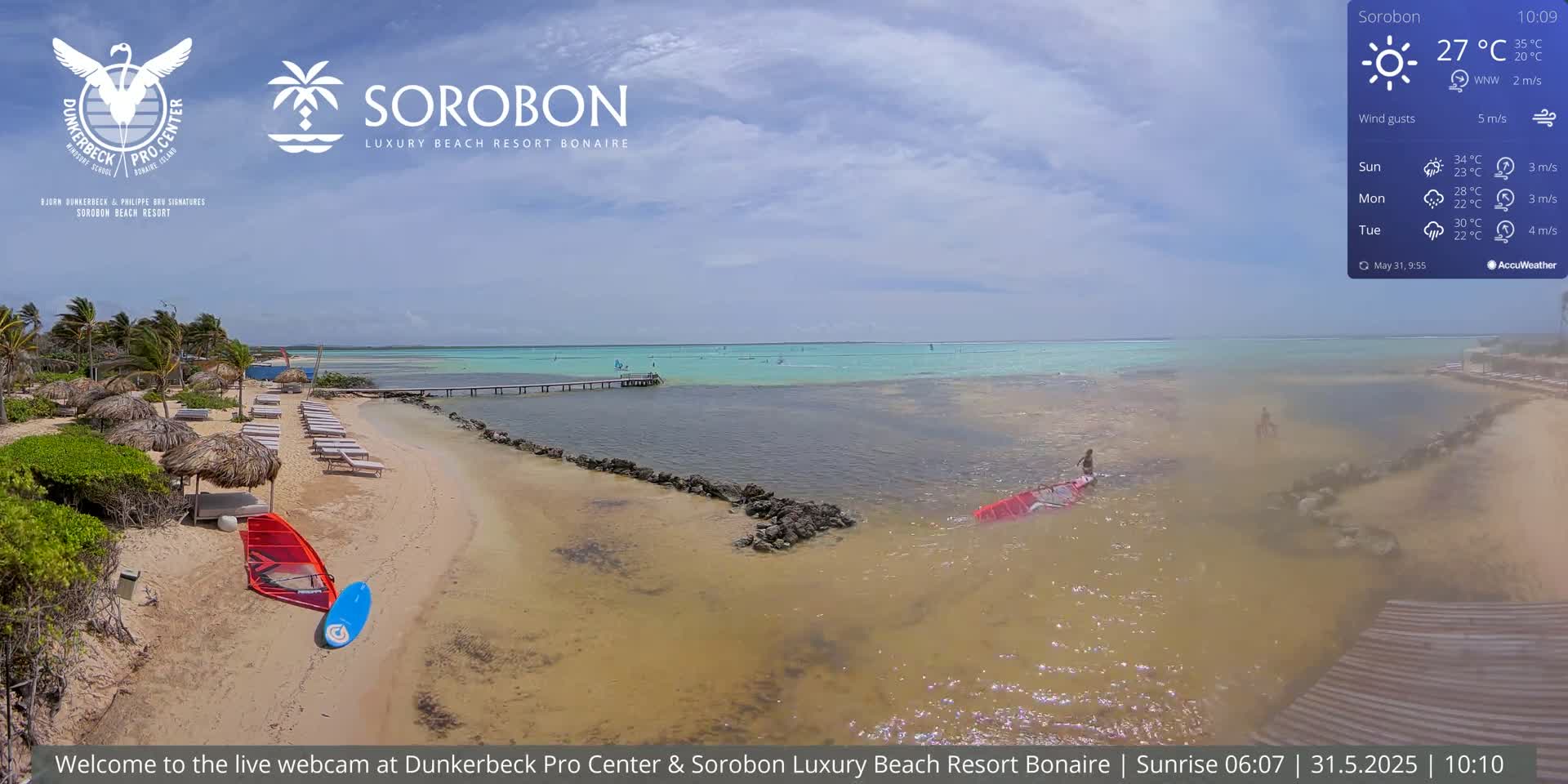 A sandy beach with lounge chairs and thatched umbrellas, leading to calm, shallow turquoise water with a few people windsurfing under a partly cloudy sky.