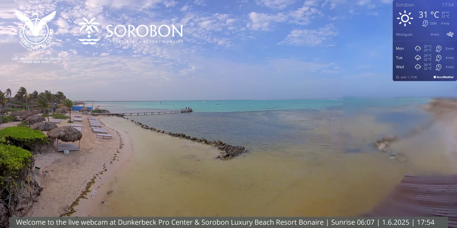 A sandy beach with lounge chairs under thatched umbrellas, a wooden pier extending into calm, turquoise water under a mostly clear sky.