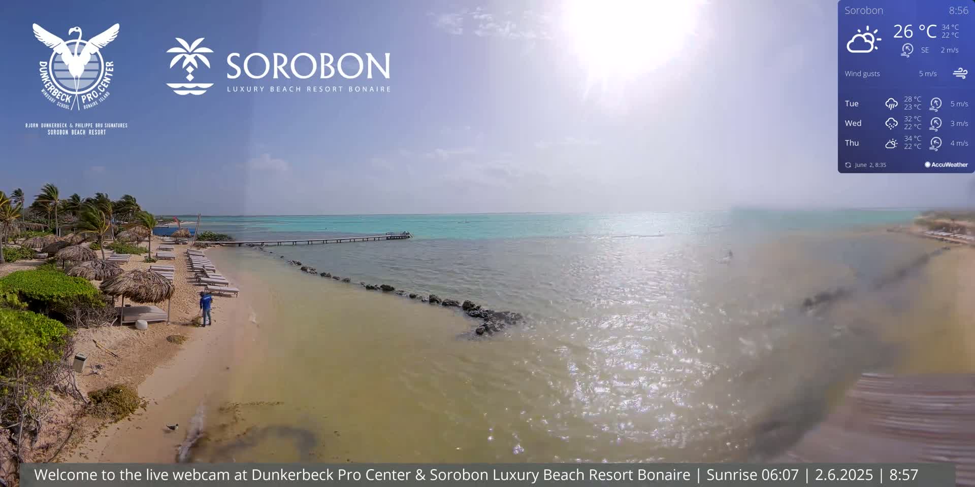 A sandy beach with lounge chairs and palm trees leads to a calm, turquoise ocean under a partly cloudy sky.