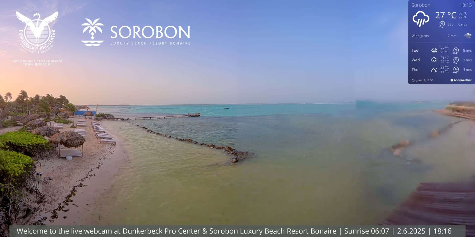A sandy beach with thatched umbrellas and lounge chairs leads to calm, turquoise water under a partly cloudy sky.