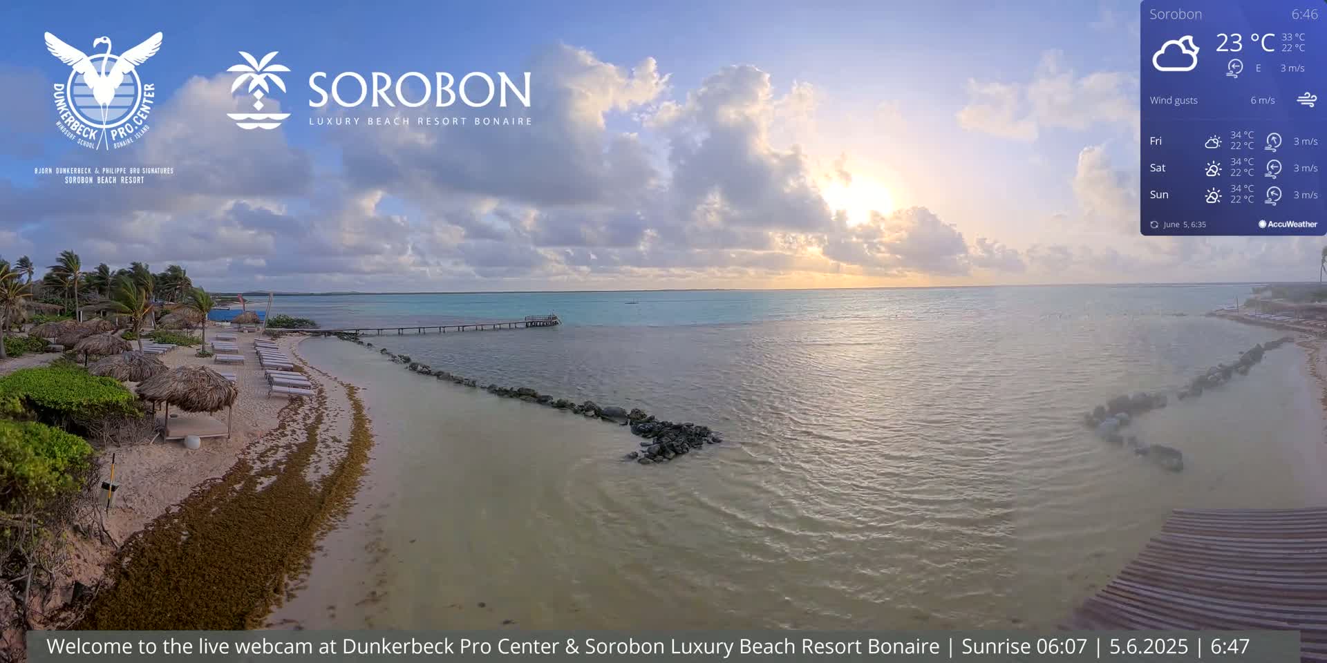 A tranquil beach scene at sunrise shows a sandy shore lined with beach umbrellas and lounge chairs, a calm sea with a pier extending into the water, and a partly cloudy sky.