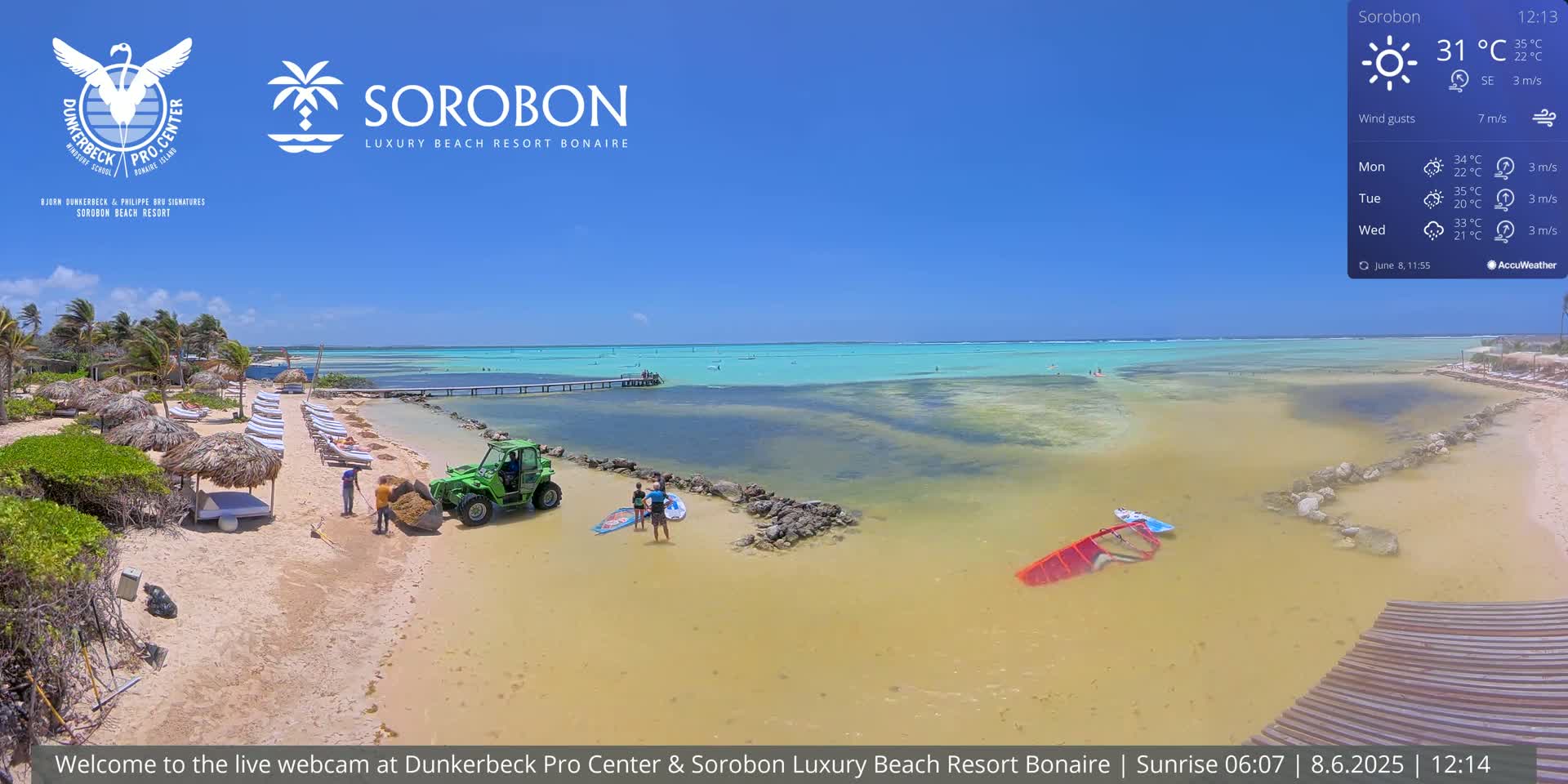 A sandy beach with lounge chairs and thatched umbrellas, a small utility vehicle, and several people near calm, shallow turquoise water under a clear blue sky.