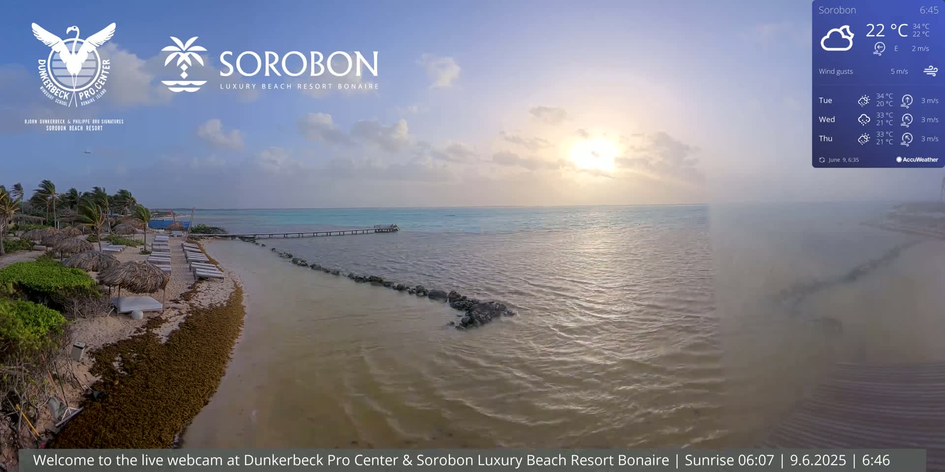 An aerial view shows a beach with lounge chairs under thatched umbrellas, a calm sea with a pier, and a partly cloudy sky with the sun rising.