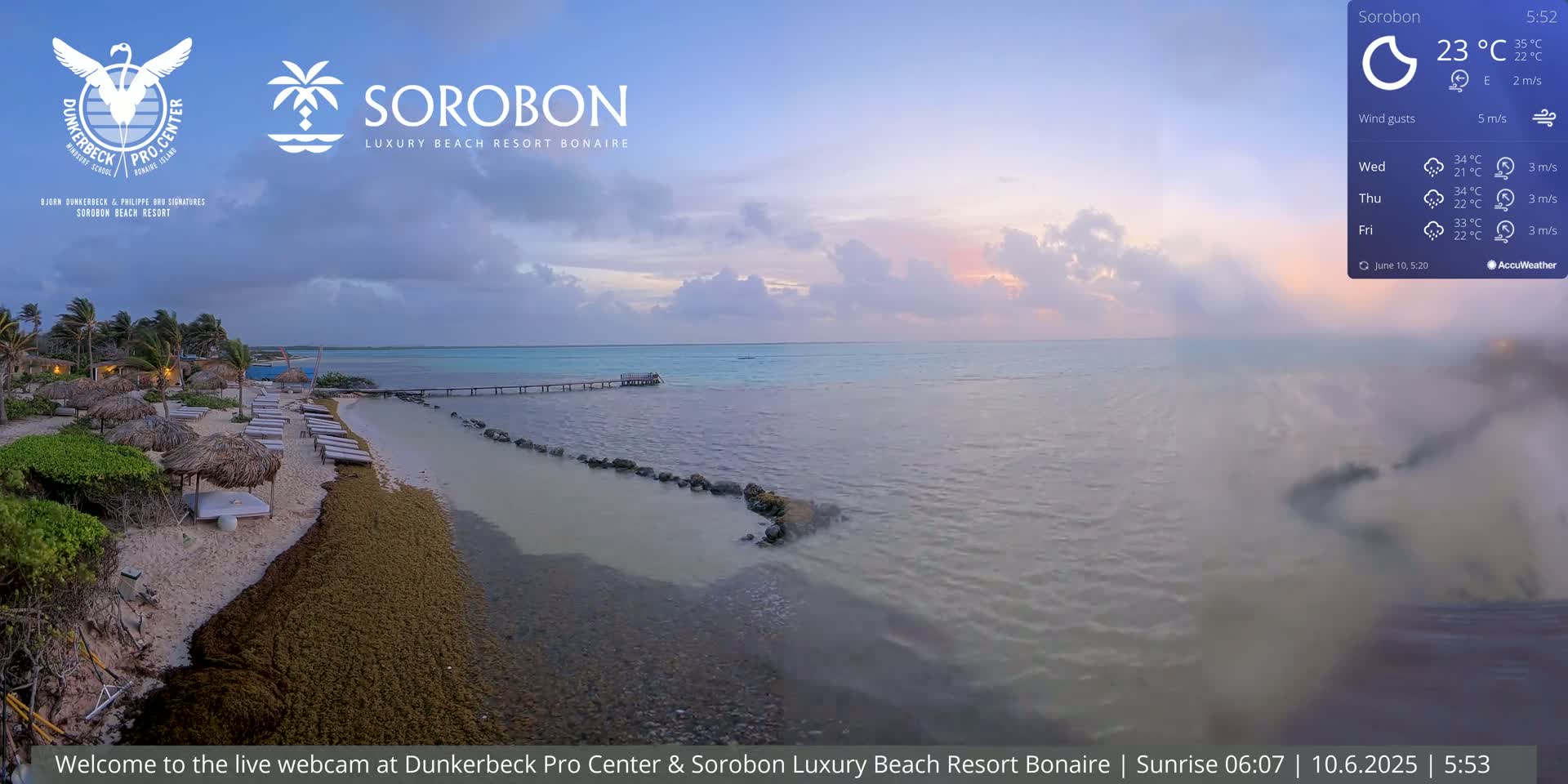 A beach with lounge chairs and thatched umbrellas under a twilight sky, overlooking a calm sea with a pier extending into the water.