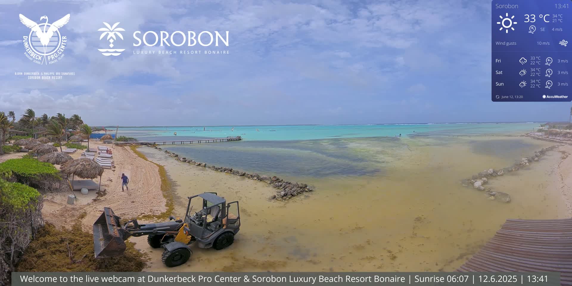 A beach with a small bobcat, lounge chairs under thatched umbrellas, and a calm, shallow turquoise ocean under a partly cloudy sky.
