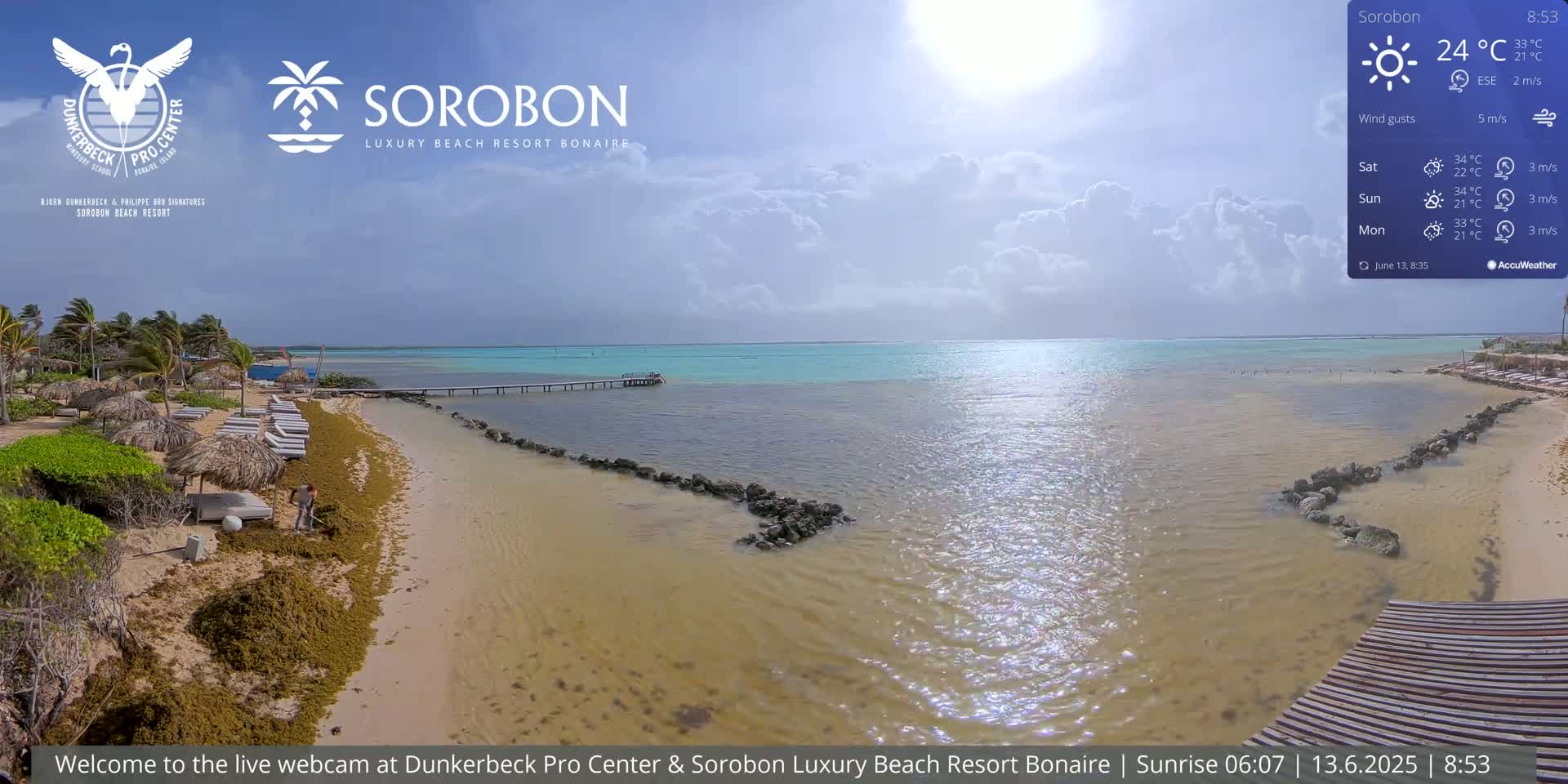 A partly cloudy day shows a sandy beach with lounge chairs, seaweed, and a person cleaning the beach, next to calm, shallow turquoise water and a wooden pier under a mostly sunny sky.