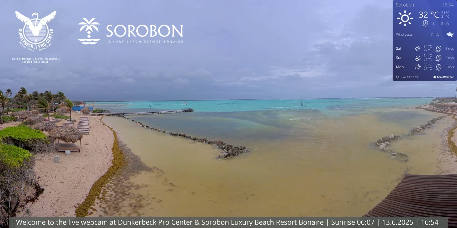 A beach with lounge chairs under thatched umbrellas, a calm turquoise sea with windsurfers, and an overcast sky.