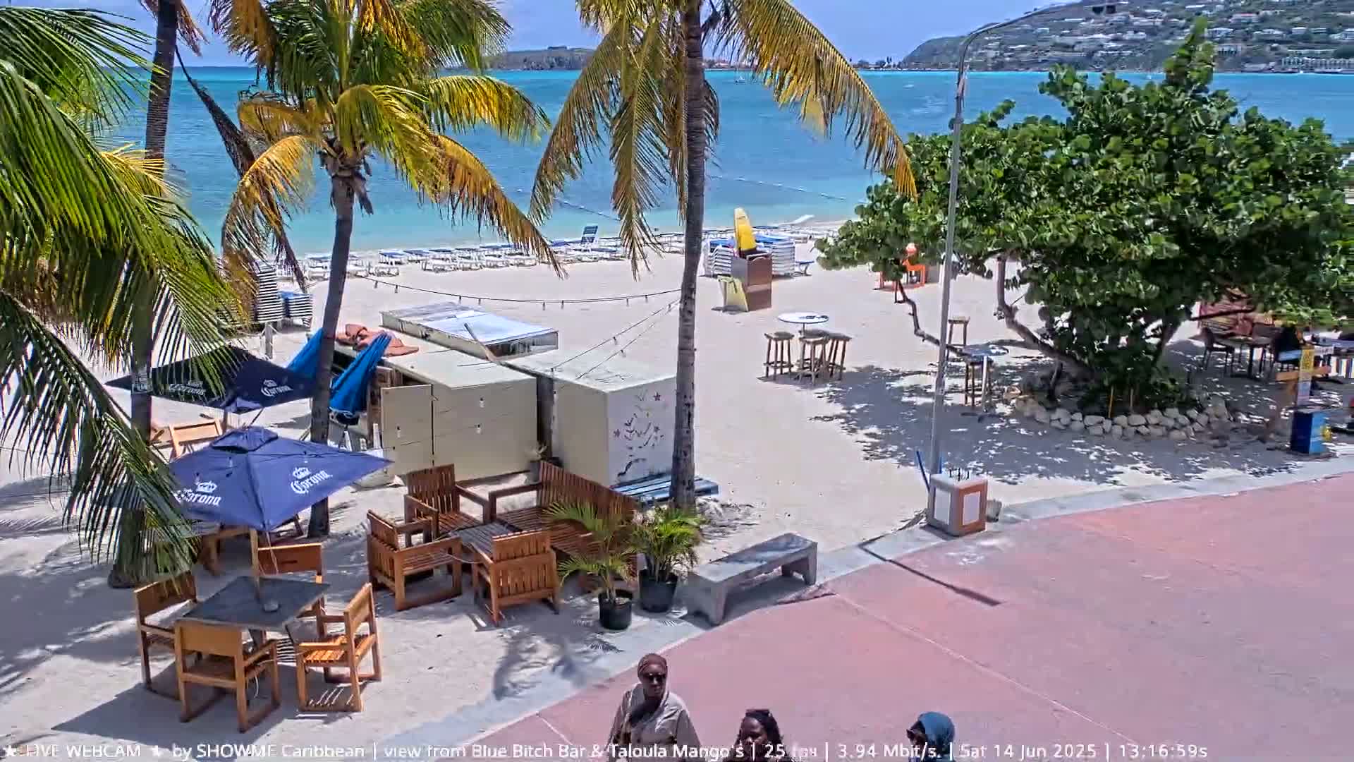 A sunny beach scene shows a sandy shore with lounge chairs, palm trees, outdoor seating areas with umbrellas, and people walking on a nearby paved walkway.