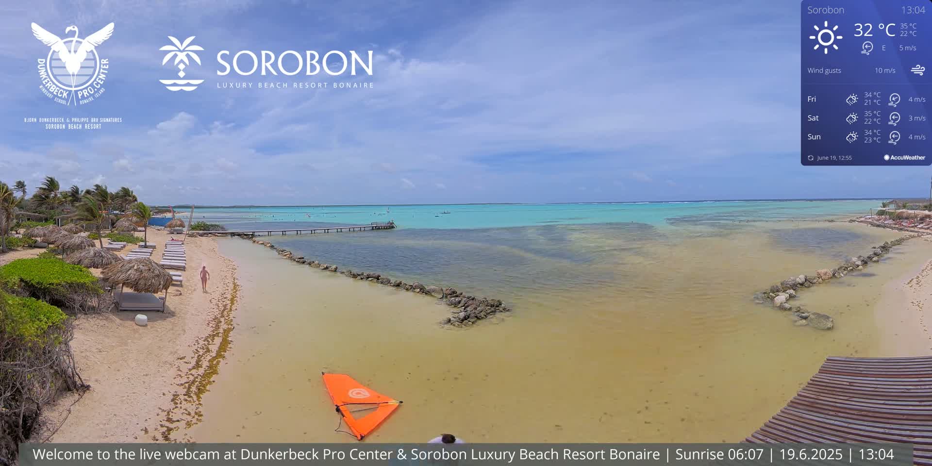 A sandy beach with lounge chairs and umbrellas leads to a calm, shallow turquoise ocean under a mostly sunny sky.