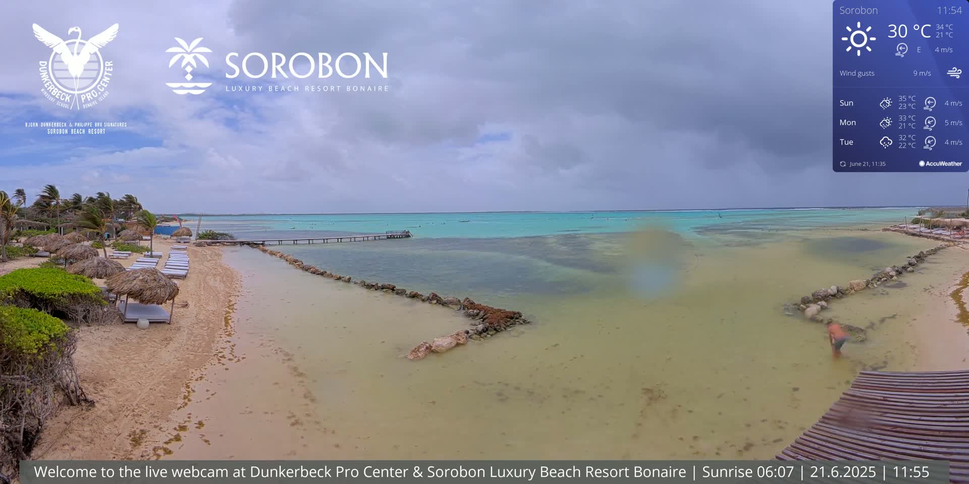 A sandy beach with thatched-roof sunshades and a calm, shallow turquoise ocean under an overcast sky.