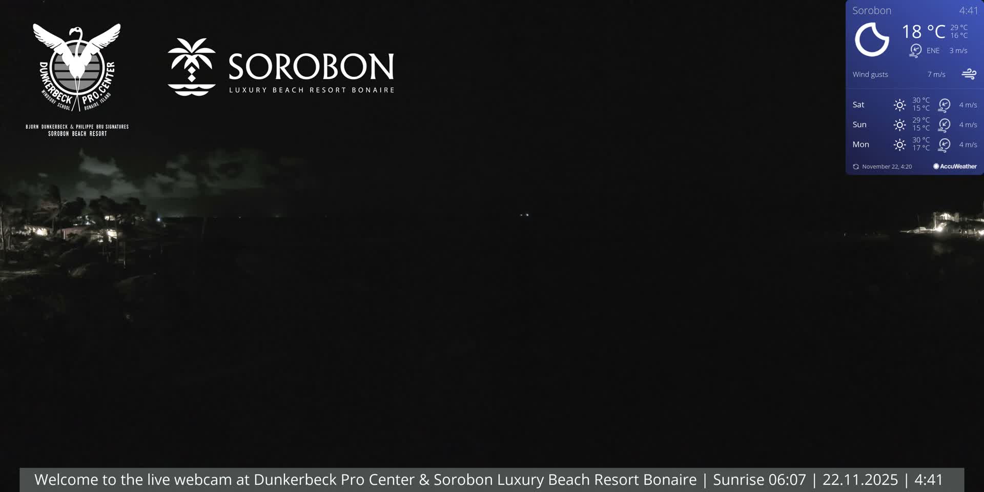 A dark, pre-dawn view of a coastline with distant resort lights reflecting on the water under a partially cloudy sky, with a temperature of 18°C.