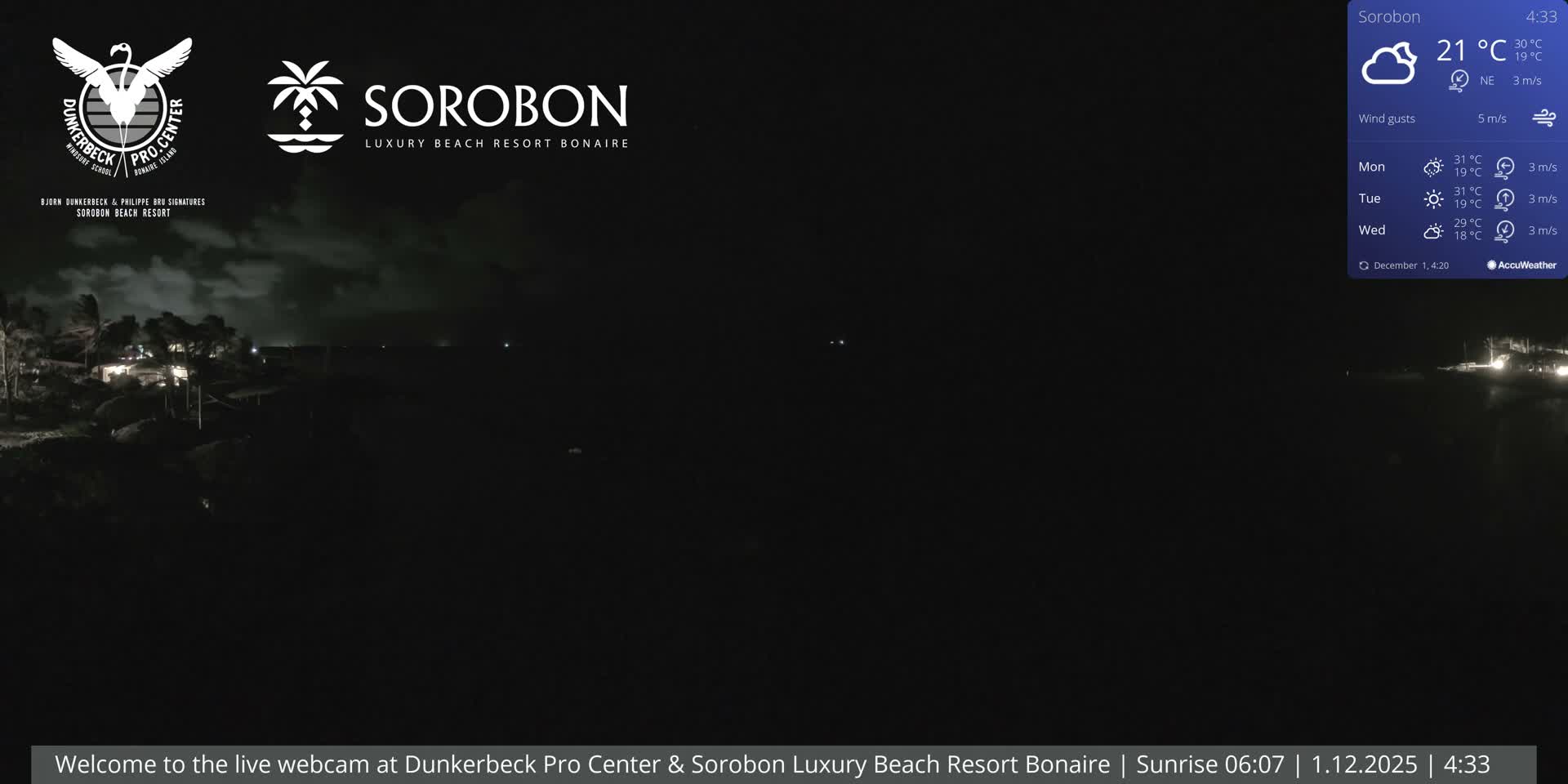 A dark nighttime view captures an illuminated coastal resort with buildings and palm trees beside the water, under cloudy skies with a temperature of 21°C and a light northeast wind.