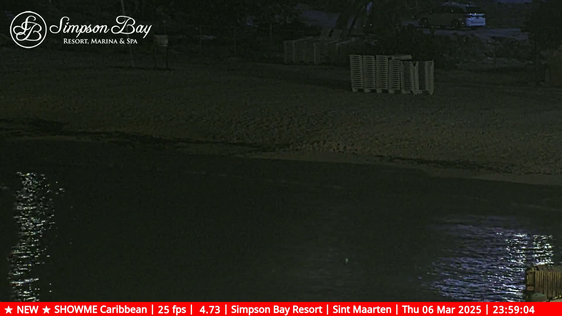 A dark, nighttime view of a sandy beach and calm water, with several folded chairs visible on the sand.