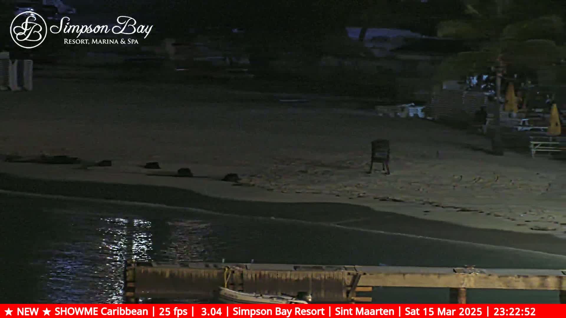 A dark, nighttime beach scene shows a mostly empty sandy shore, calm water lapping at the edge, and a small boat moored at a wooden dock.