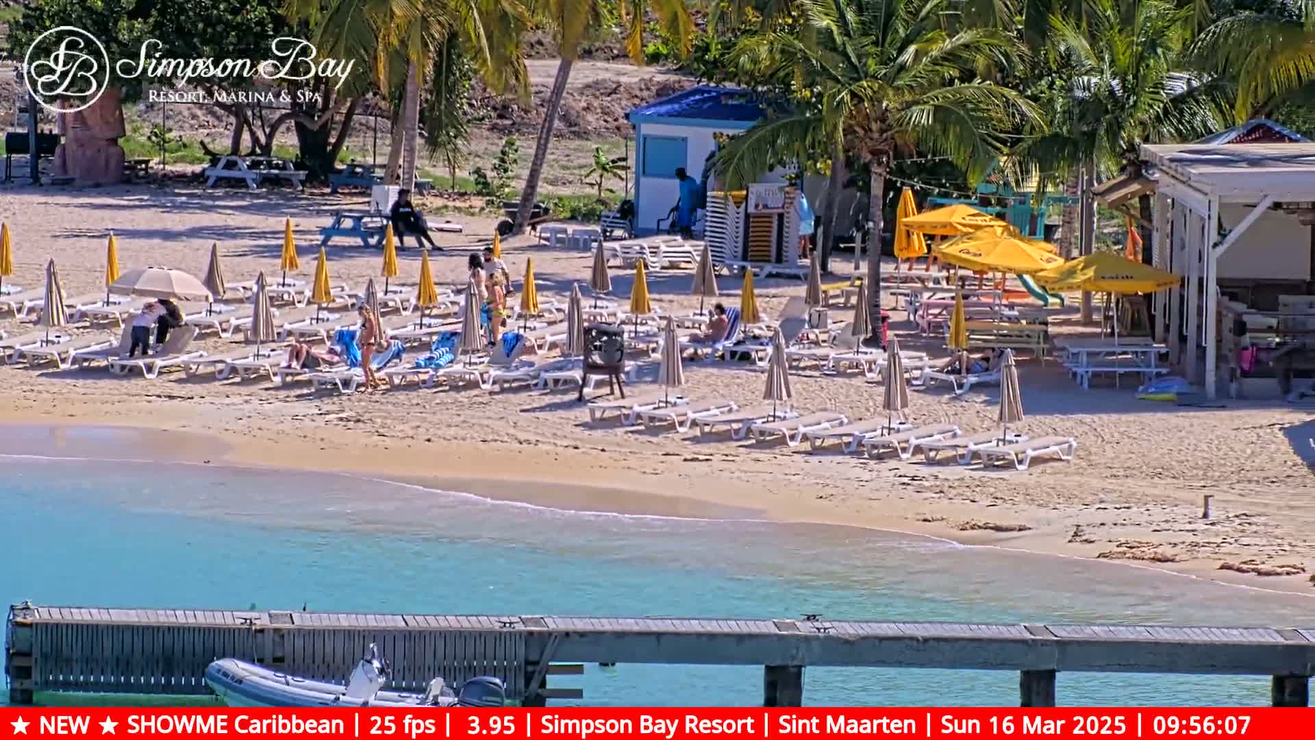 A sandy beach with numerous lounge chairs and umbrellas under a sunny sky, adjacent to a small building and calm turquoise water.