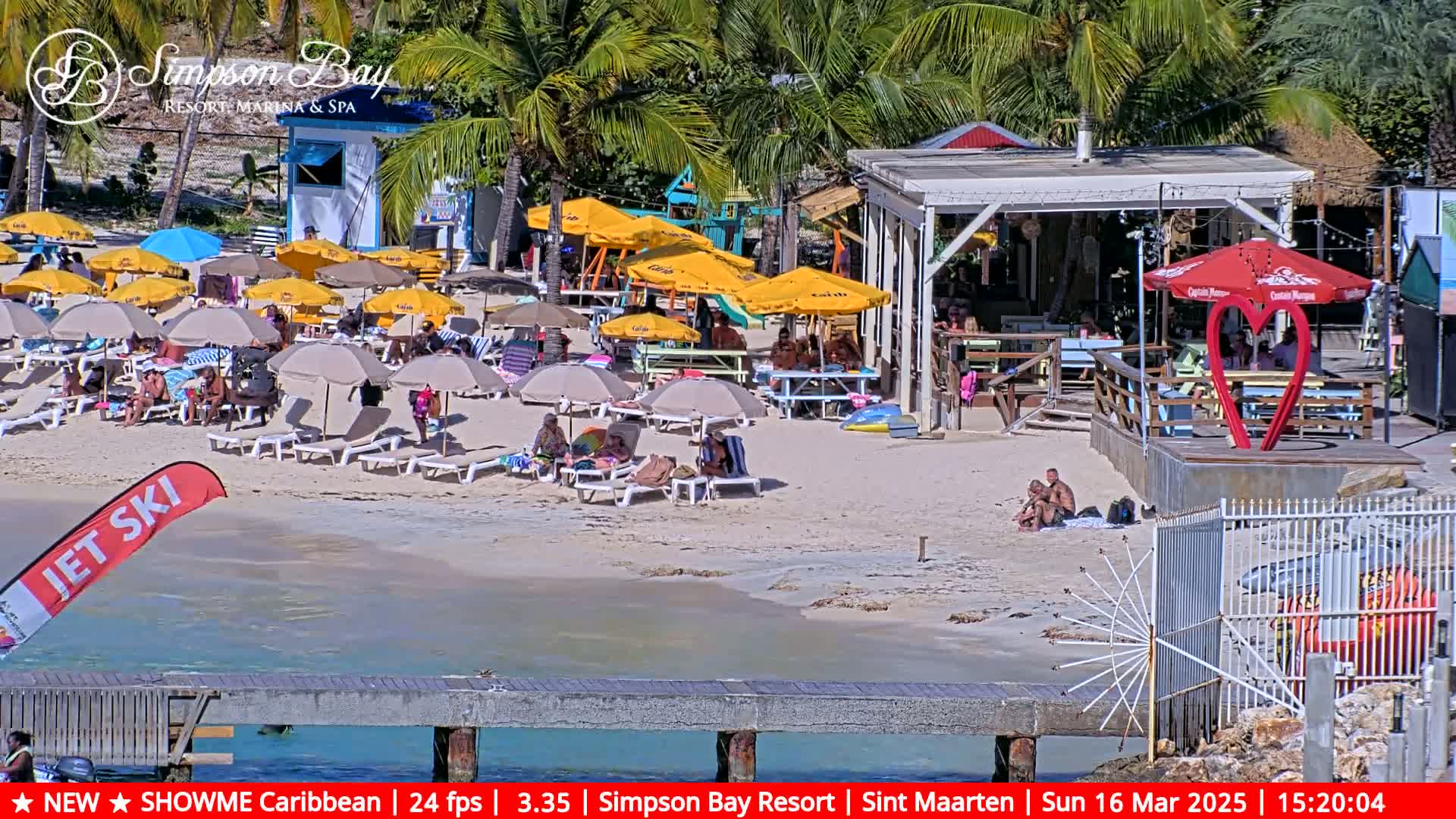 A sunny beach scene shows numerous beach umbrellas, lounge chairs, and people relaxing on a sandy beach near a beachfront bar and restaurant.