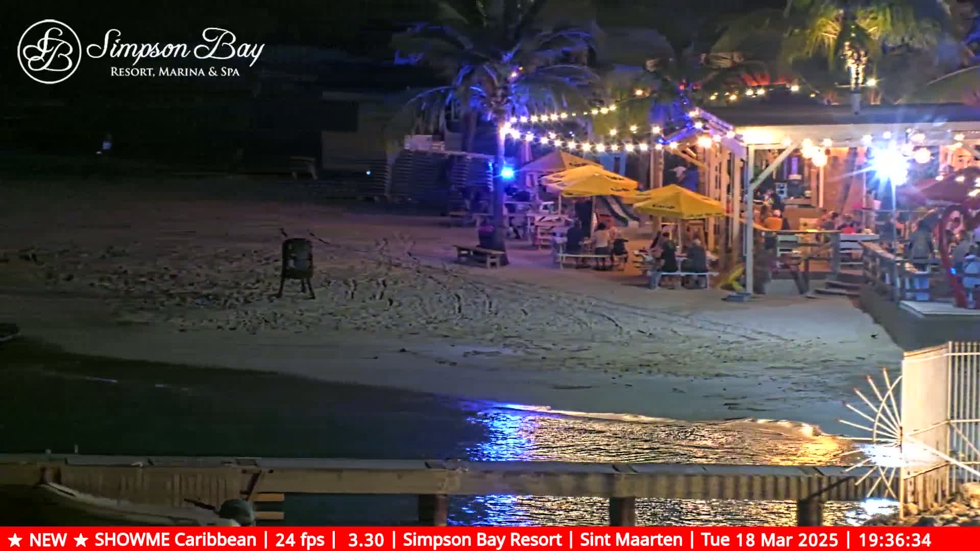 A nighttime beach scene shows a beachfront restaurant with patrons seated at tables under string lights, illuminated by the lights and the reflection of lights on the water.