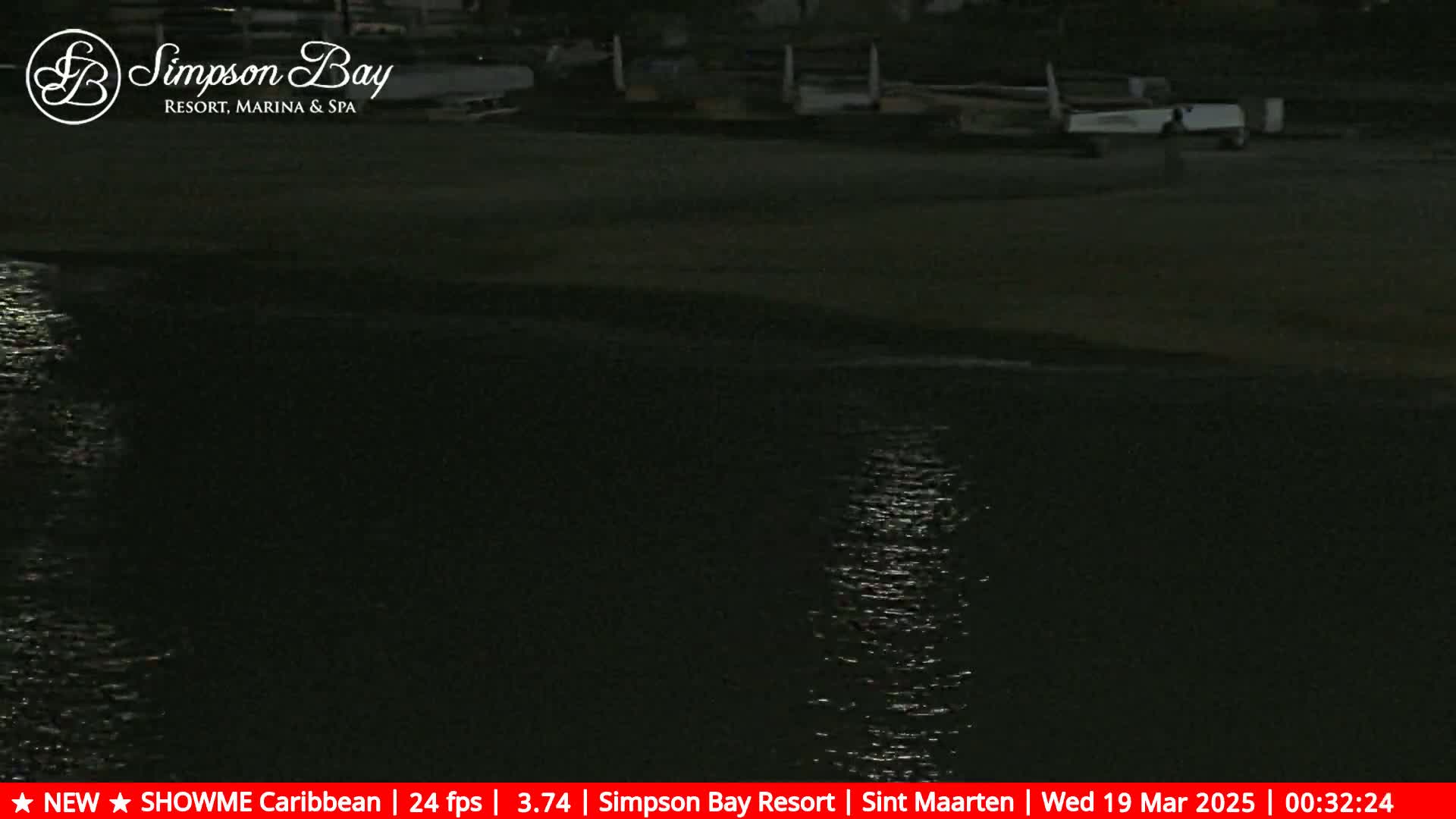 A dark, nighttime view of calm water reflecting light, with a shoreline and several boats parked nearby.
