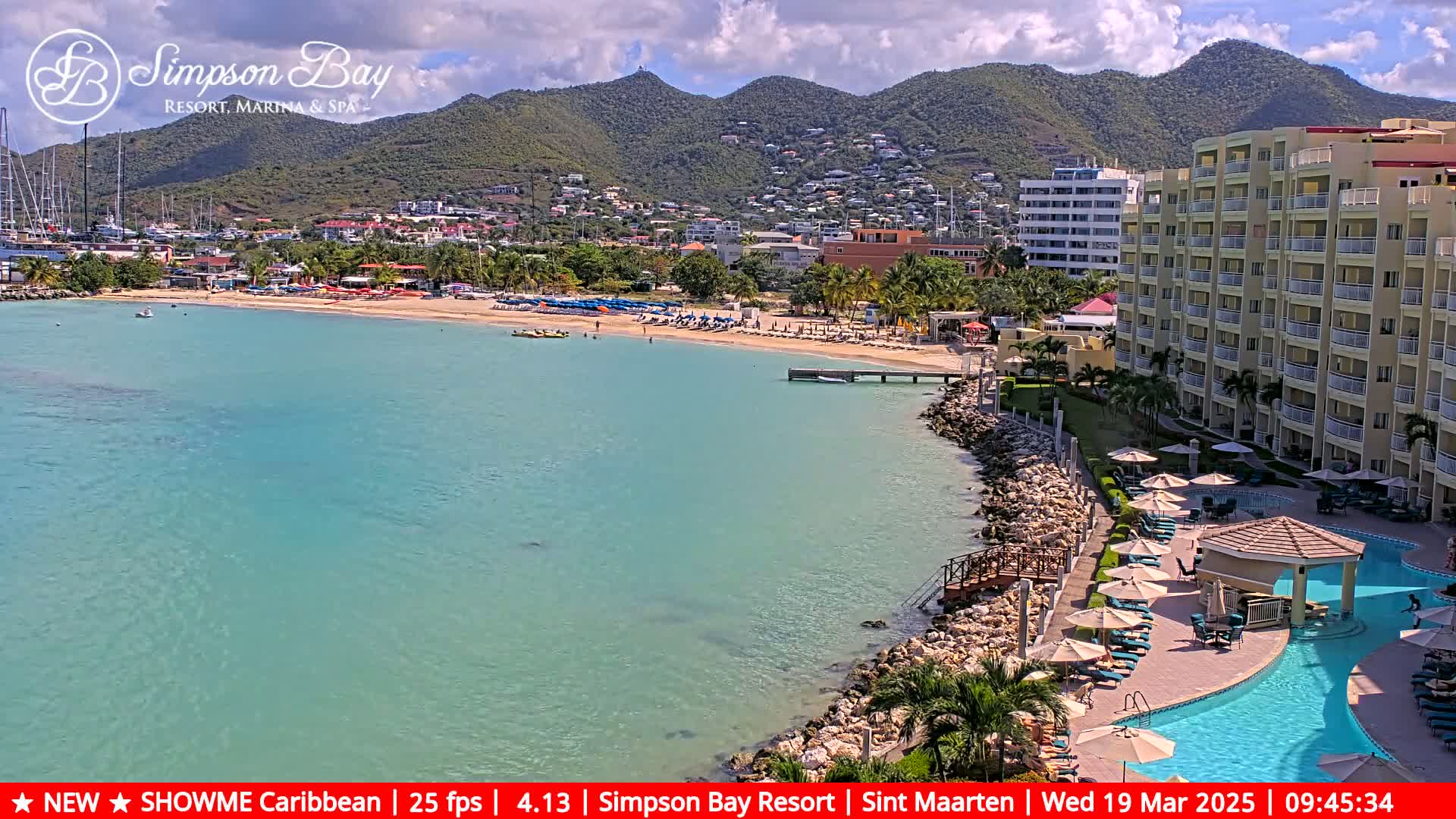 A partly cloudy day shows a tropical beach with turquoise water, a resort with a pool, and mountains in the background.