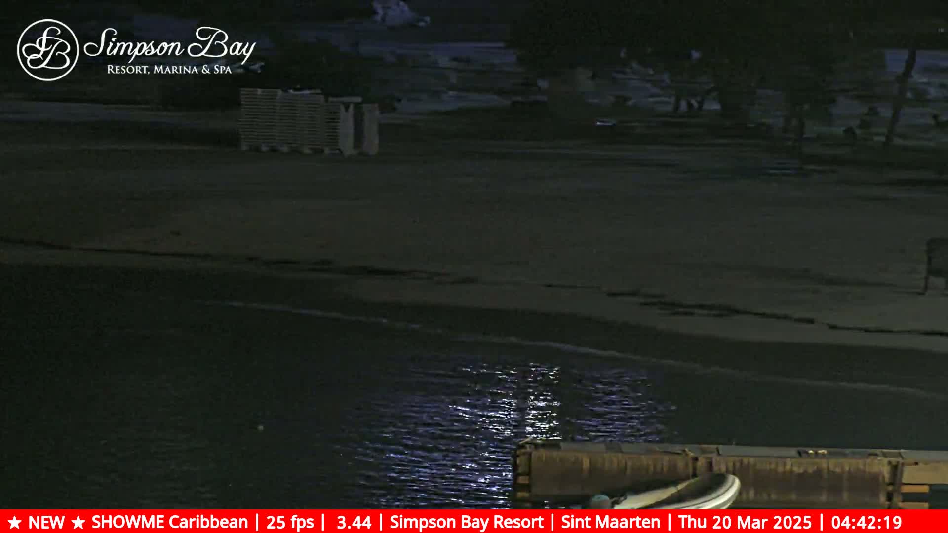 A dark nighttime beach scene shows a calm body of water lapping at the shore near a dock, with some beach furniture visible in the background.