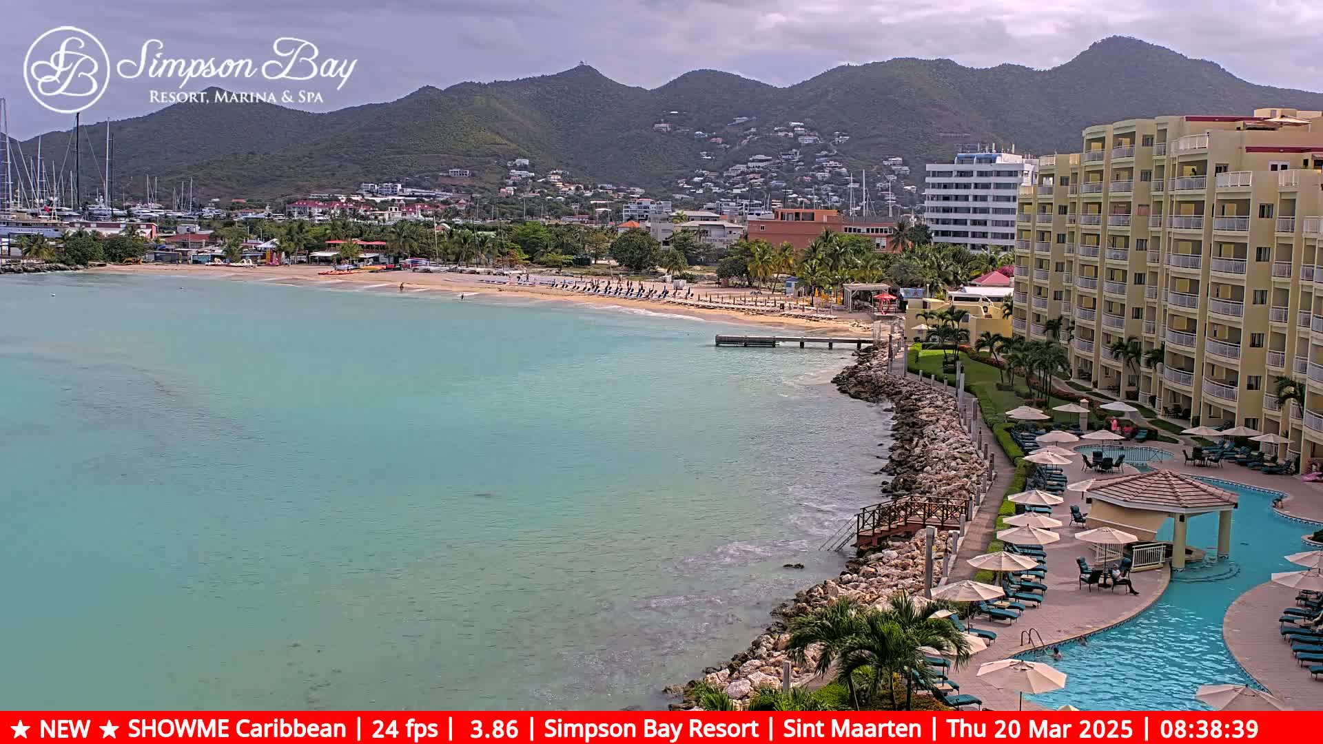 A tranquil beach with turquoise water is seen next to a resort with a swimming pool, all set against a backdrop of verdant mountains under a partly cloudy sky.